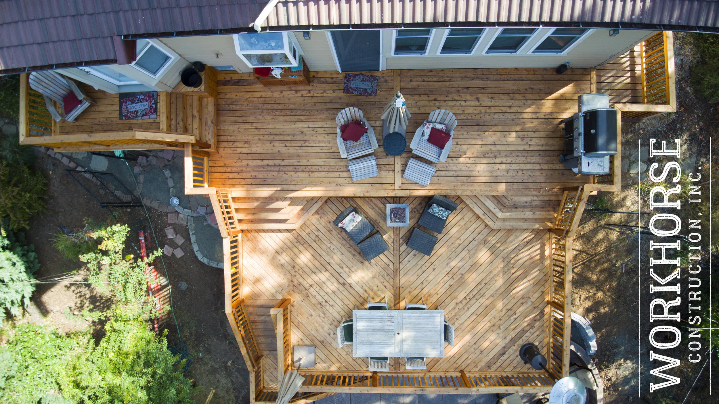 Top-down view of a wooden deck attached to a house with outdoor furniture including chairs, a table with an umbrella, and a BBQ grill, surrounded by greenery and garden landscaping.
