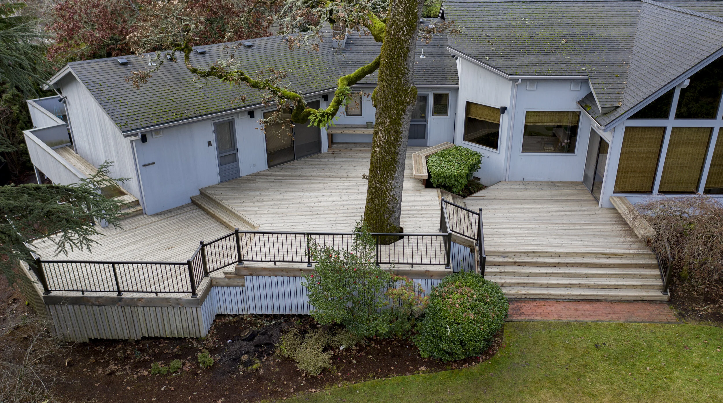 Aerial view of a backyard with a large wooden deck, a tree growing through the deck, and a house with a gray roof and white exterior walls.