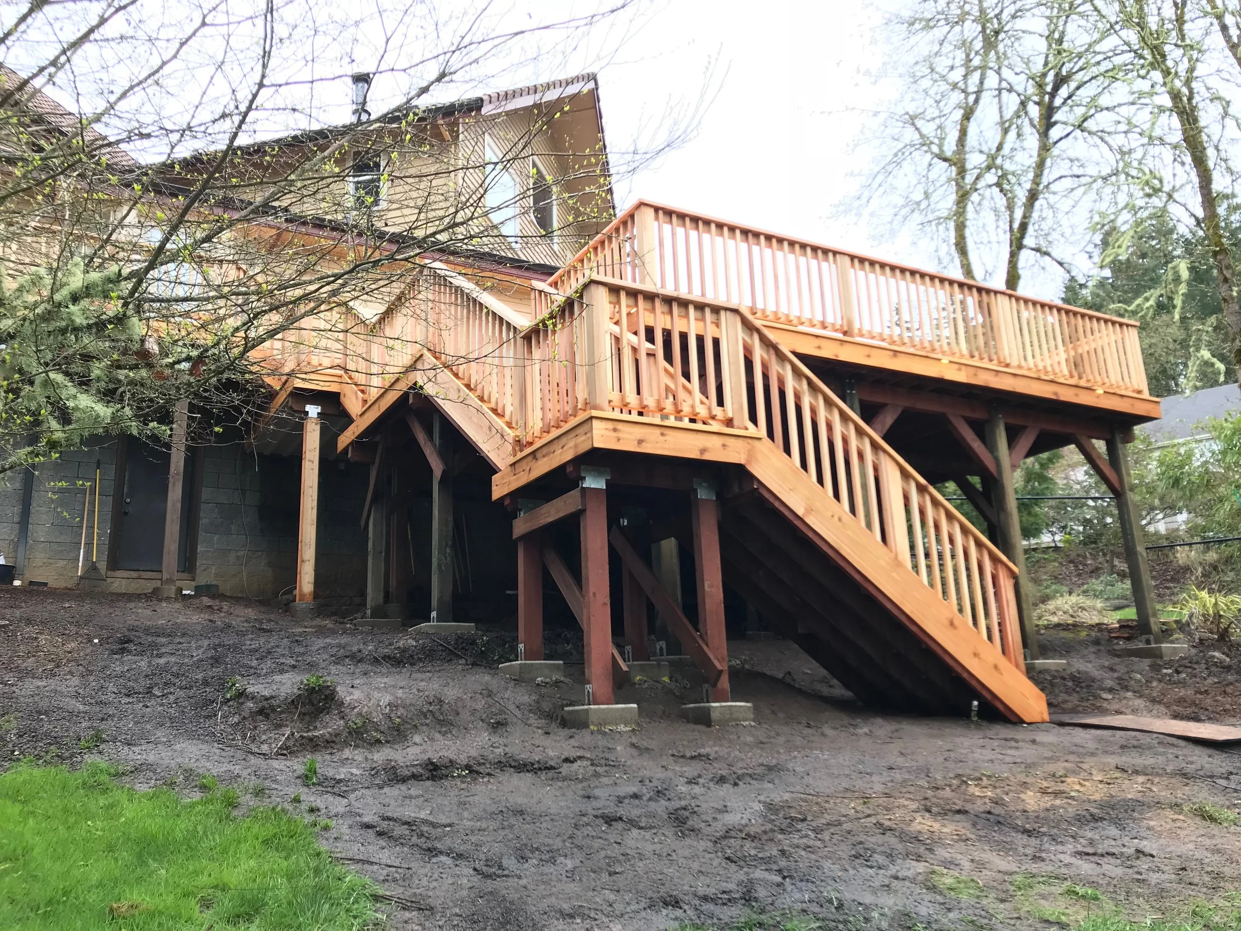 A house with a large wooden deck and stairs. The house is surrounded by trees, some with bare branches, and the yard is partly covered with green grass and dark dirt.