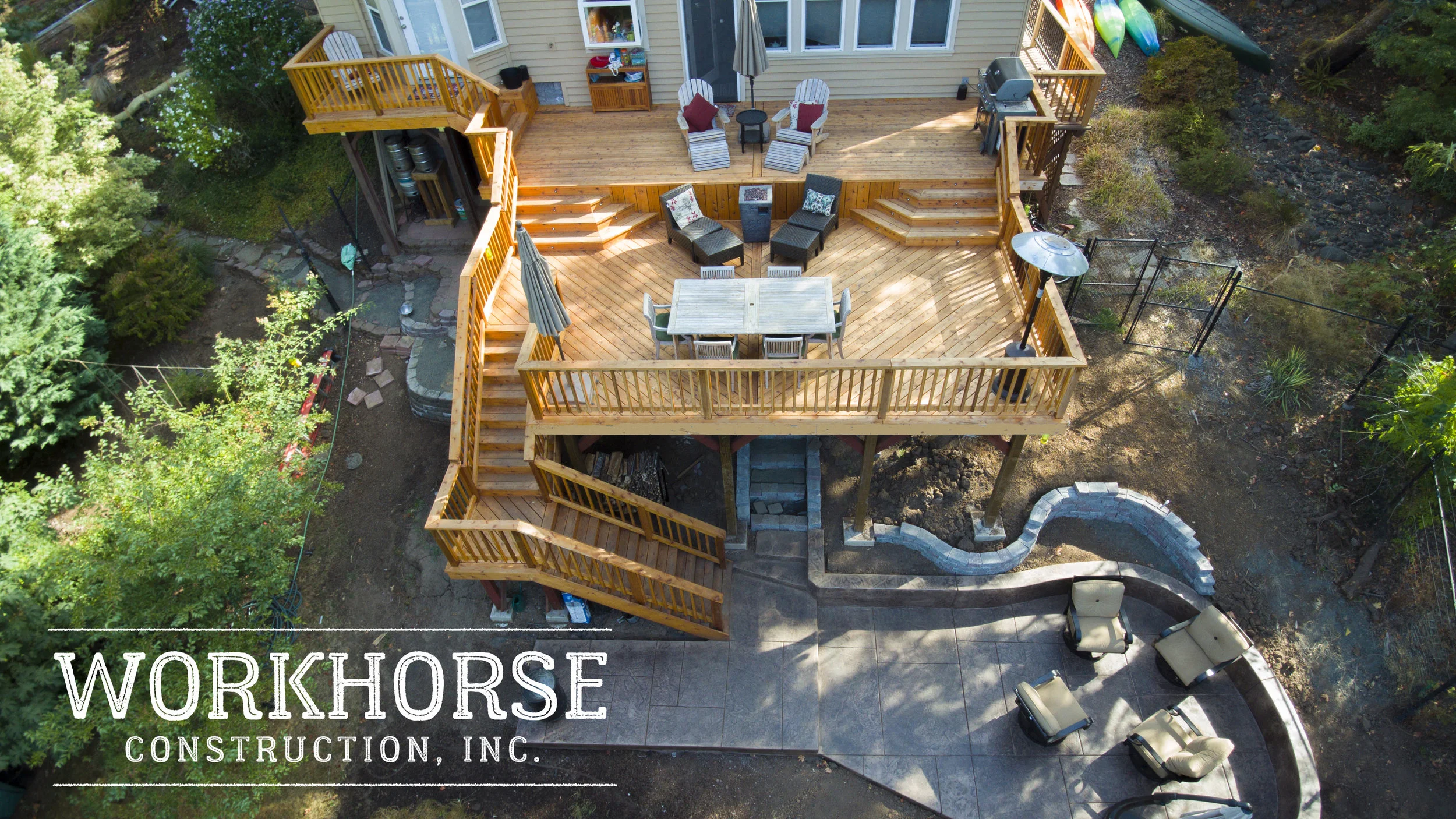 A large wooden multi-level deck attached to a yellow house with stairs, seating areas, and a barbecue grill, surrounded by greenery and trees.