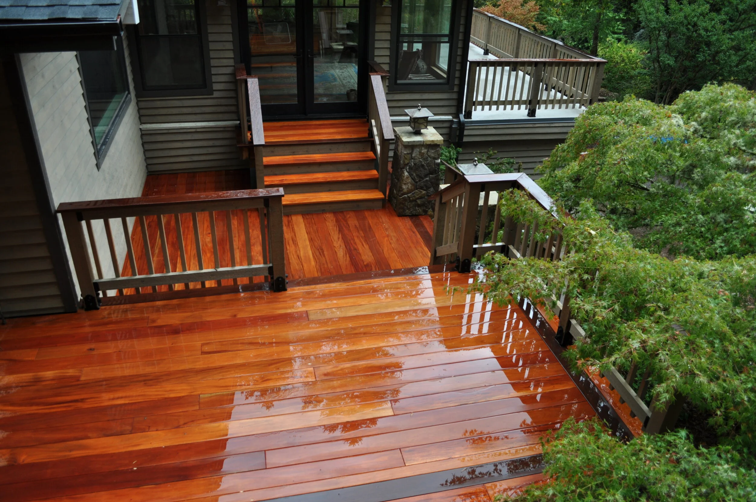 A wooden deck attached to a house with sliding glass doors. The deck has railings and is surrounded by lush green trees. The surface of the deck is shiny and appears wet.