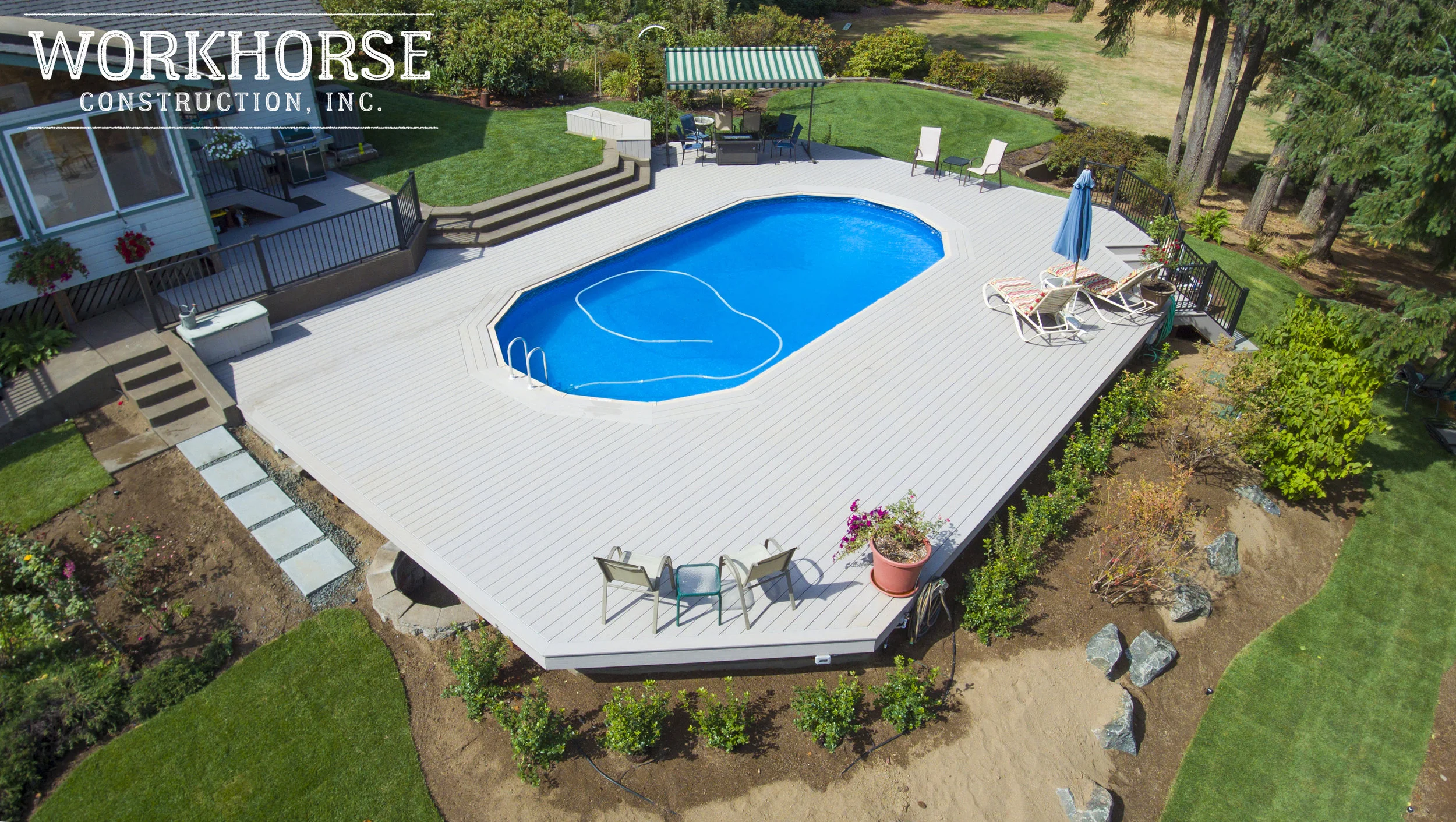 A backyard with a swimming pool surrounded by a white deck, outdoor furniture, and landscaped garden with trees and plants.