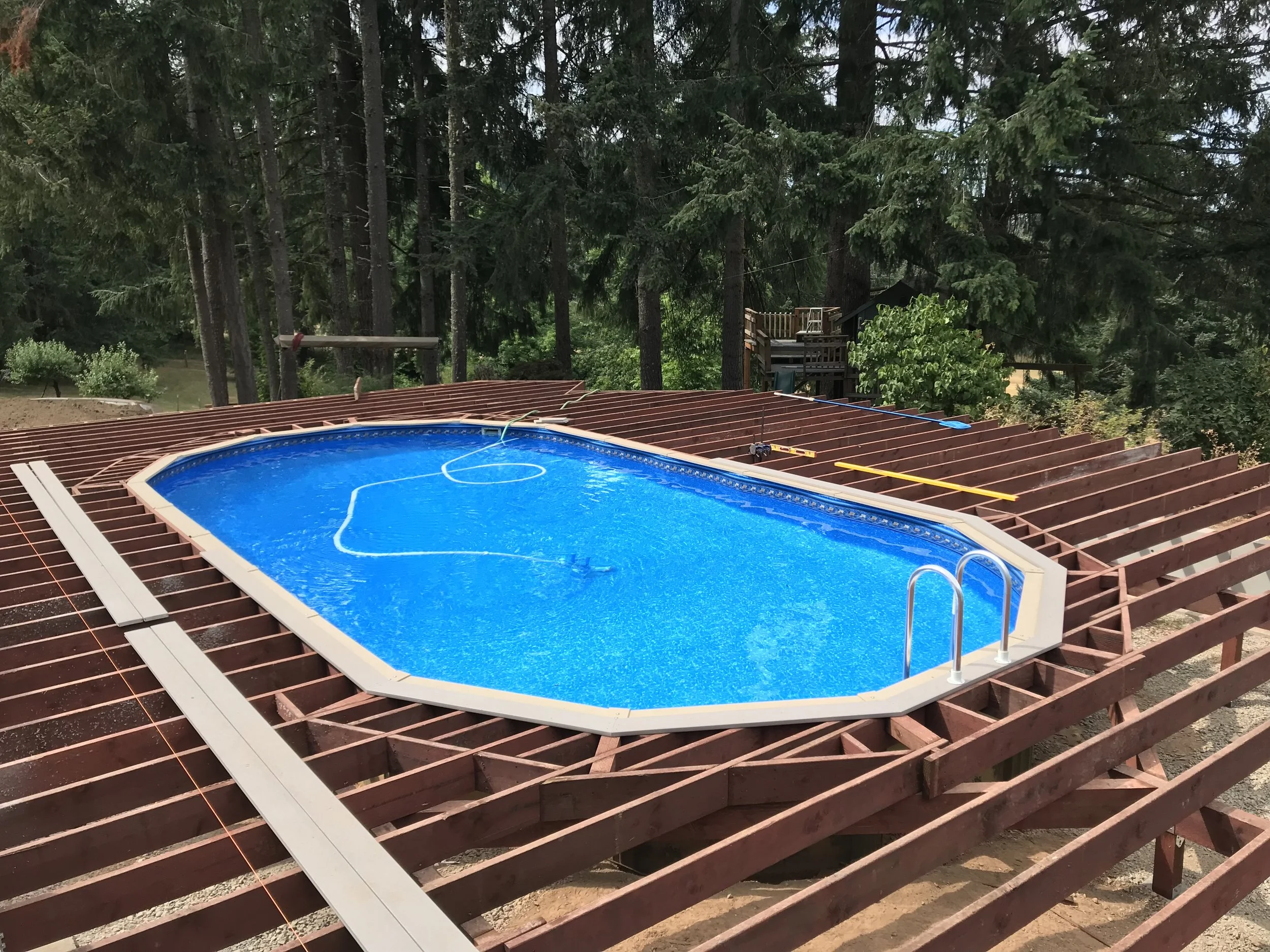 A swimming pool with a blue liner under construction, surrounded by a wooden deck framework in a forested backyard.