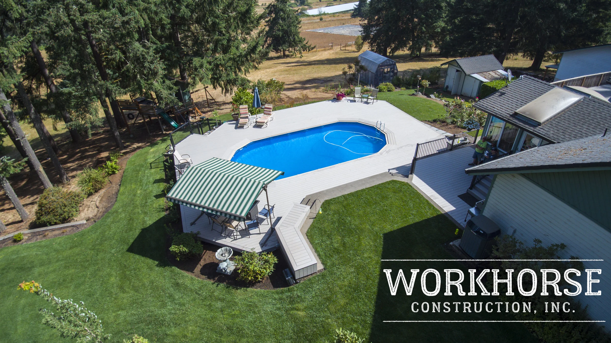 A residential backyard features a kidney-shaped swimming pool surrounded by a white deck with lounge chairs, a small seating area with a table and chairs, and a striped awning. The yard is well-manicured with green grass, plants, and tall pine trees.