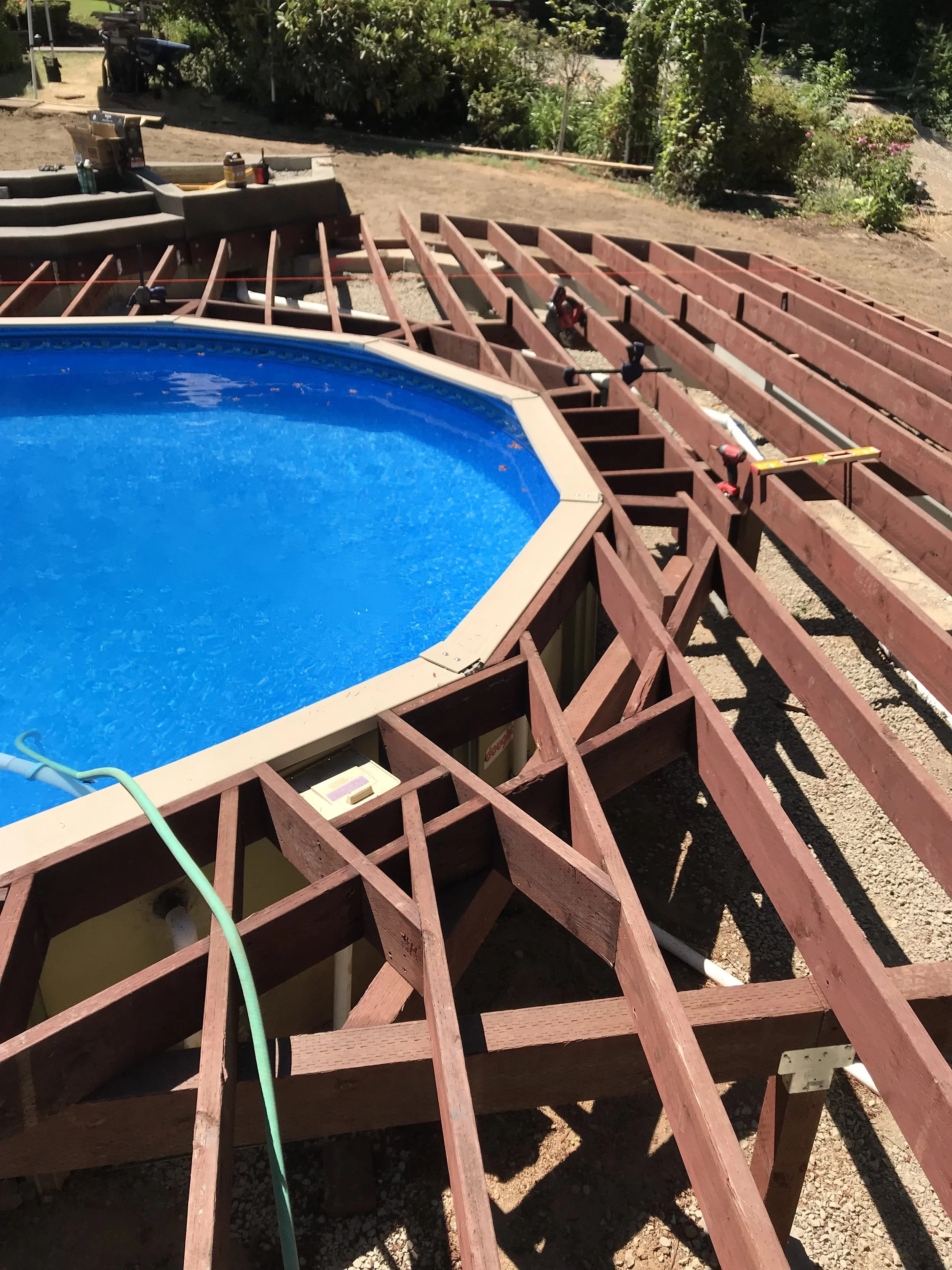 Wooden pool deck under construction with an above-ground swimming pool filled with water, tools, and supplies on the partially built deck.