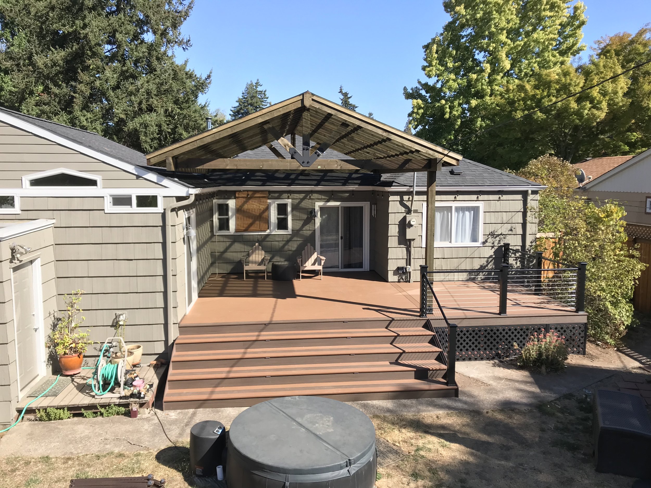 Backyard with wooden deck, potted plants, and unfinished roof extension on house surrounded by tall trees.