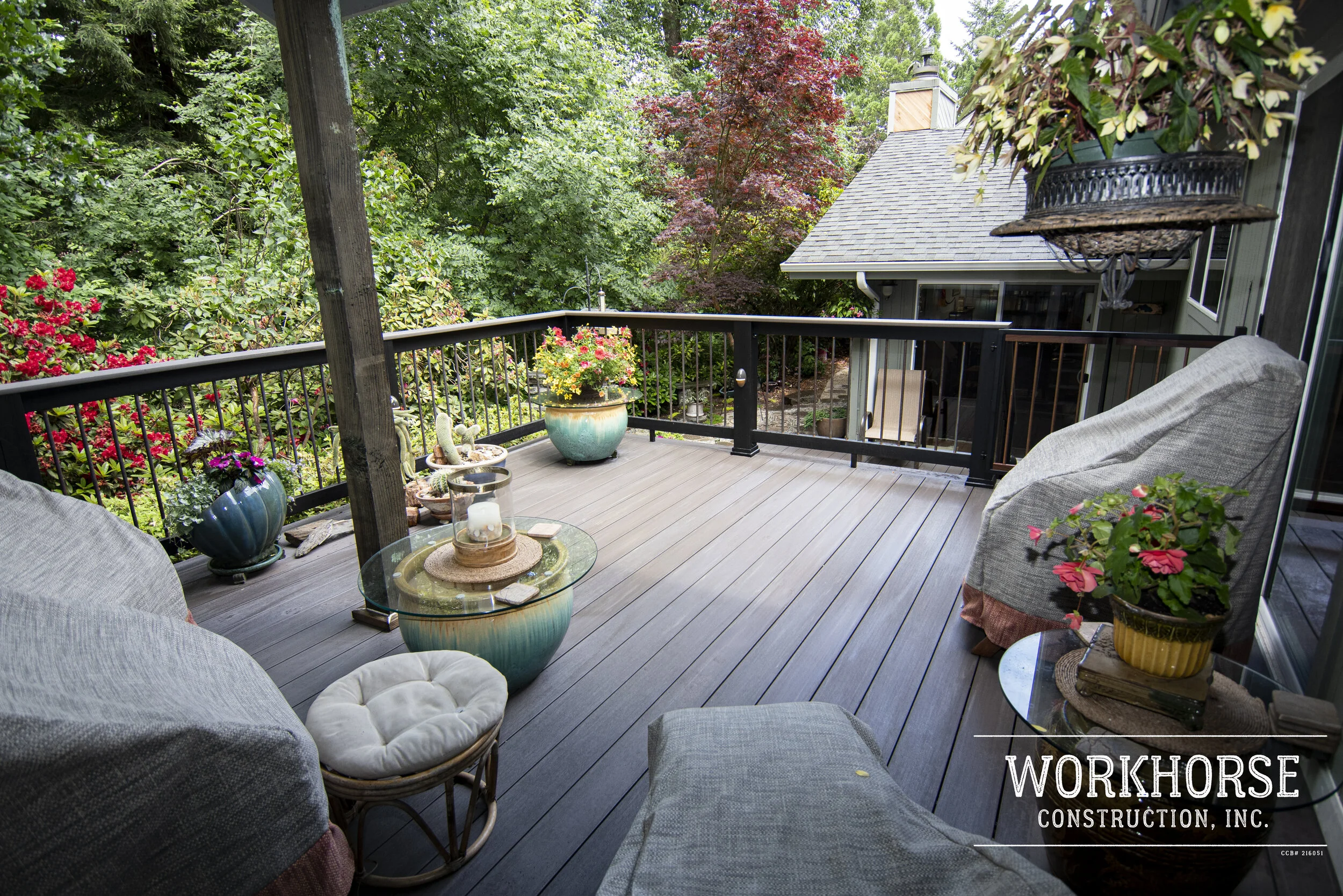 Balcony with outdoor furniture and potted plants, overlooking a lush green garden with trees and house rooftops in the background.
