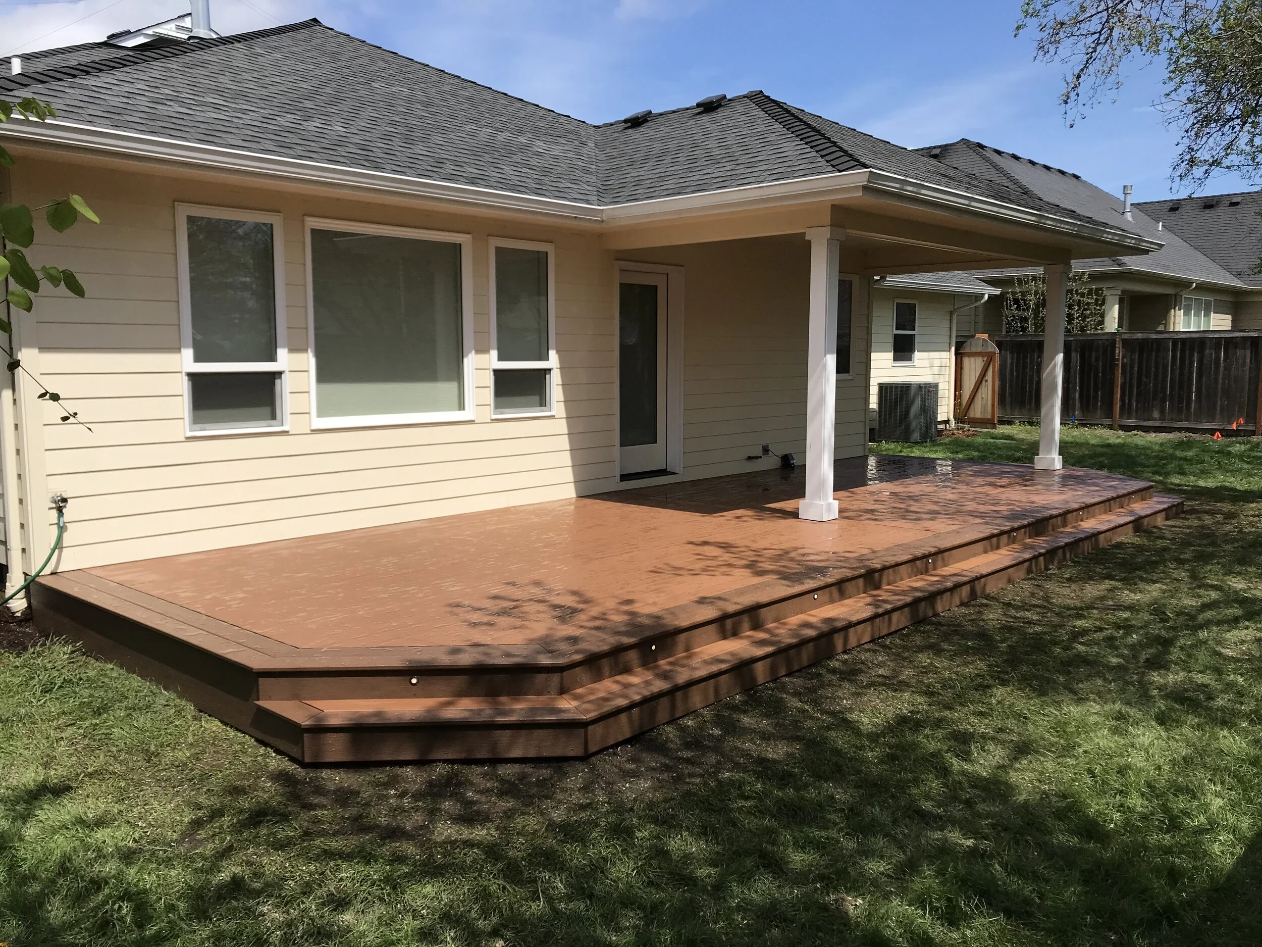 A backyard patio with a wood deck attached to a house with beige siding and multiple windows, surrounded by a grassy yard and fenced in backyard.