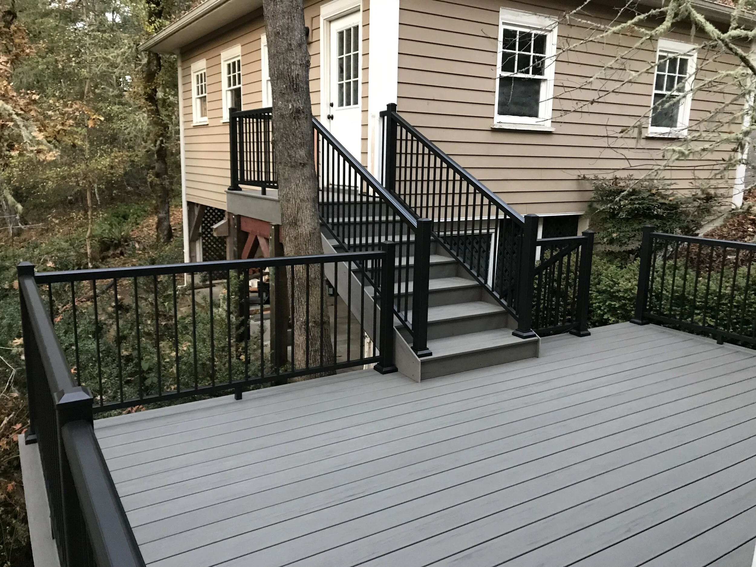 A beige house with white trim and a black railing staircase leading to a white door, surrounded by trees with autumn leaves and a deck in the foreground.