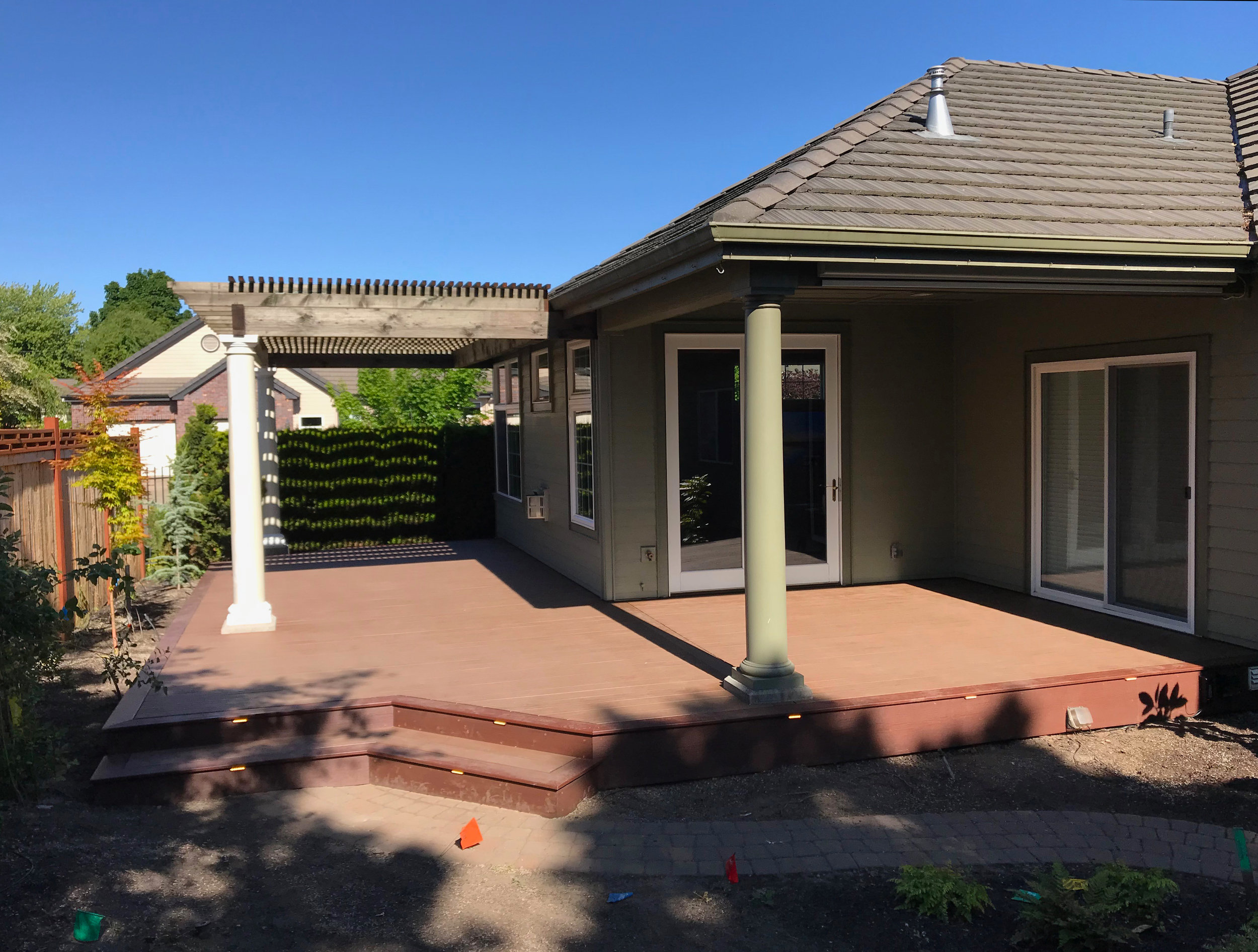 Backyard patio with a wooden deck, supported by a green column, attached to a house with sliding glass doors, surrounded by a garden with trees and bushes, under a clear blue sky.