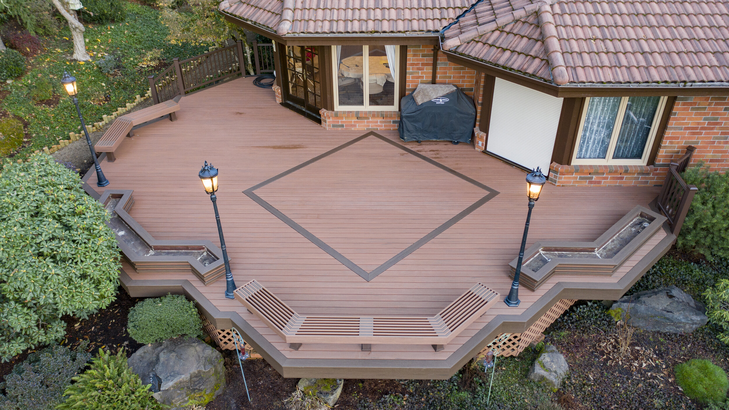 A wooden deck with built-in benches and decorative trim, surrounded by greenery, with three lampposts lit. The deck extends from a brick house with sliding glass doors and an asphalt shingle roof, and has a fenced garden area.