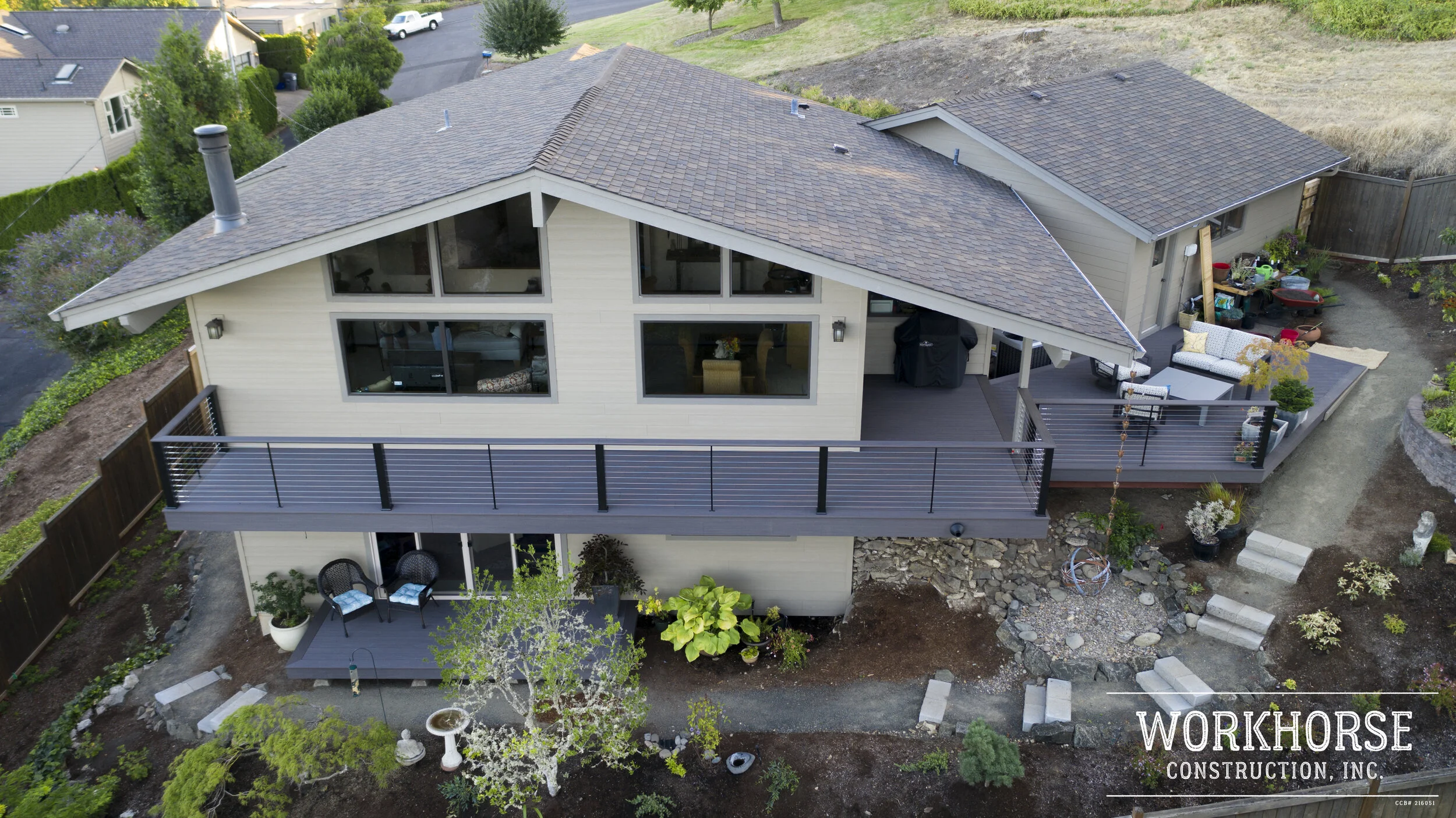 An aerial view of a modern two-story house with a large balcony and outdoor seating area. The house has a gray roof with multiple slopes, beige siding, and large windows. The yard surrounding the house has garden beds, trees, and landscaping in progr