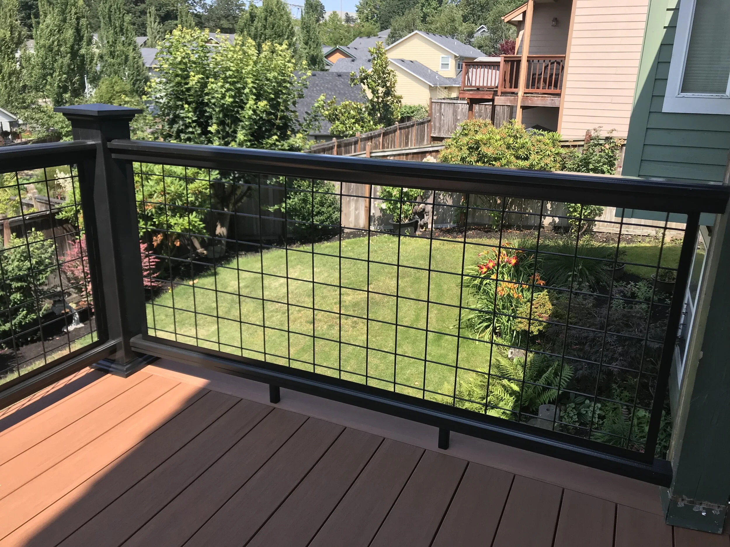 View from a balcony showing a backyard with green grass, various trees, and shrubs, enclosed by a wooden and wire fence. Neighboring houses with various roof colors are visible in the background.