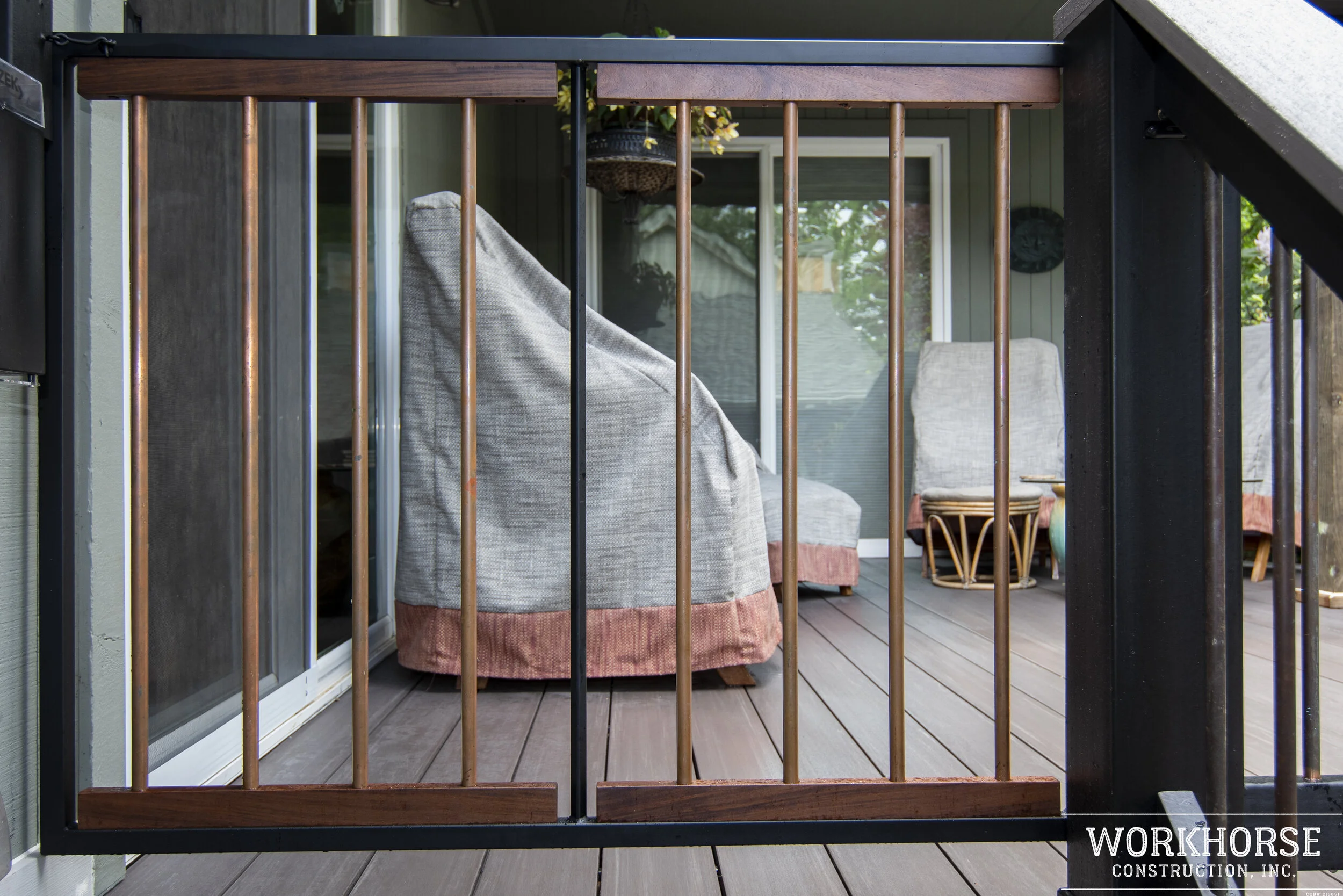 Close-up of a balcony railing with wooden and metal bars, overlooking a porch with chairs and potted plants outside a house.