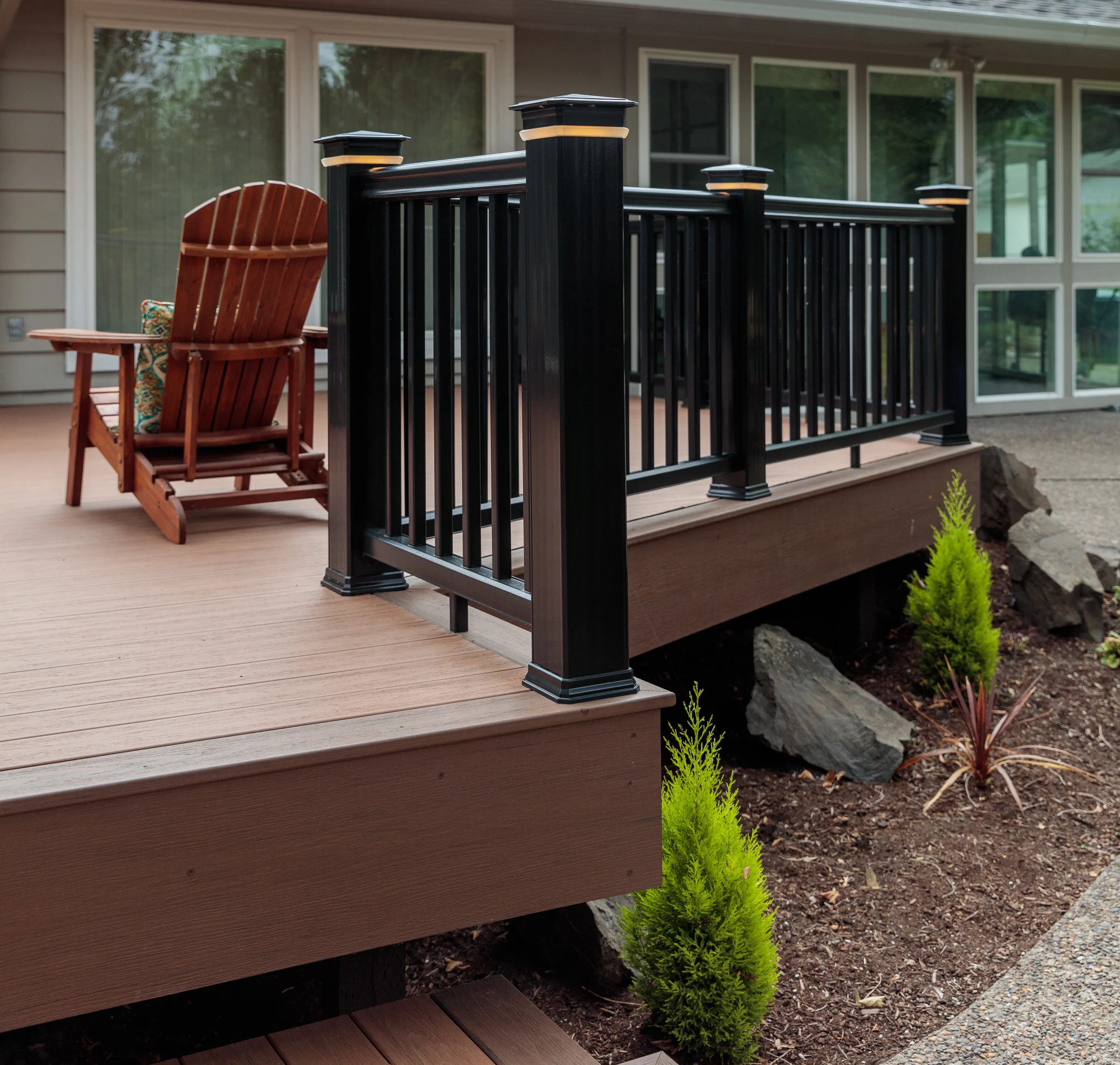A wooden Adirondack chair and a small wooden rocking chair on a deck, with a black metal railing and a house with large windows in the background, surrounded by garden plants and rocks.