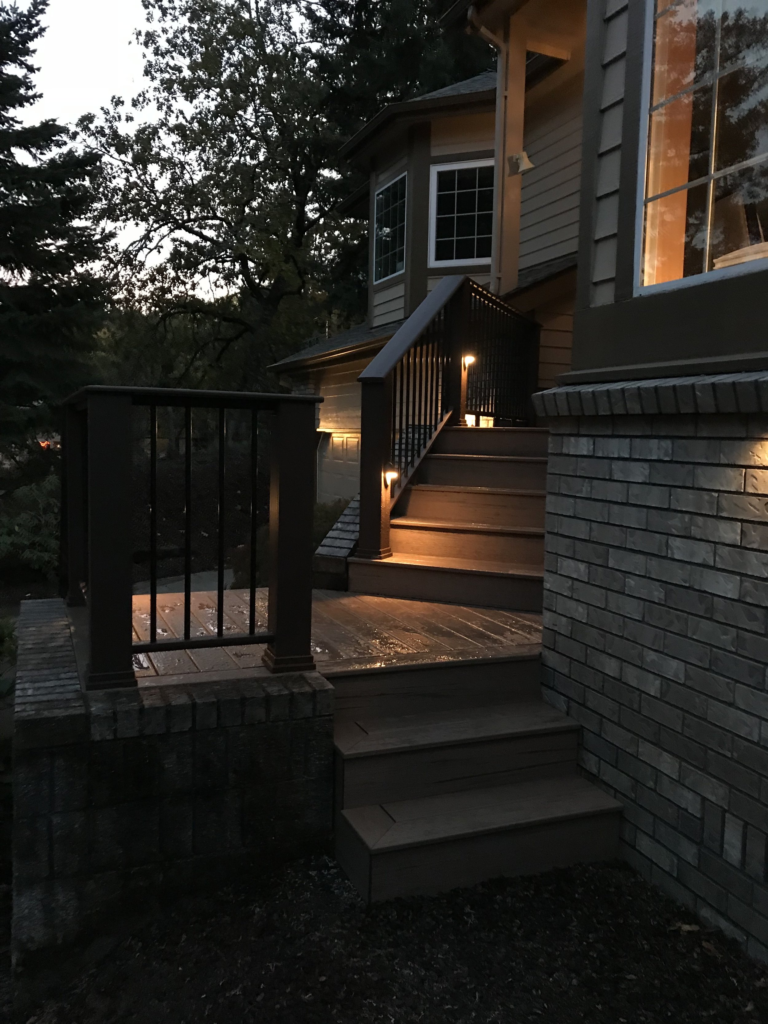 A house with a brick and siding exterior, illuminated by outdoor lights near a small staircase. The house has a large window with a warm glow, and a tree with leaves is visible in the background, indicating dusk or early evening.