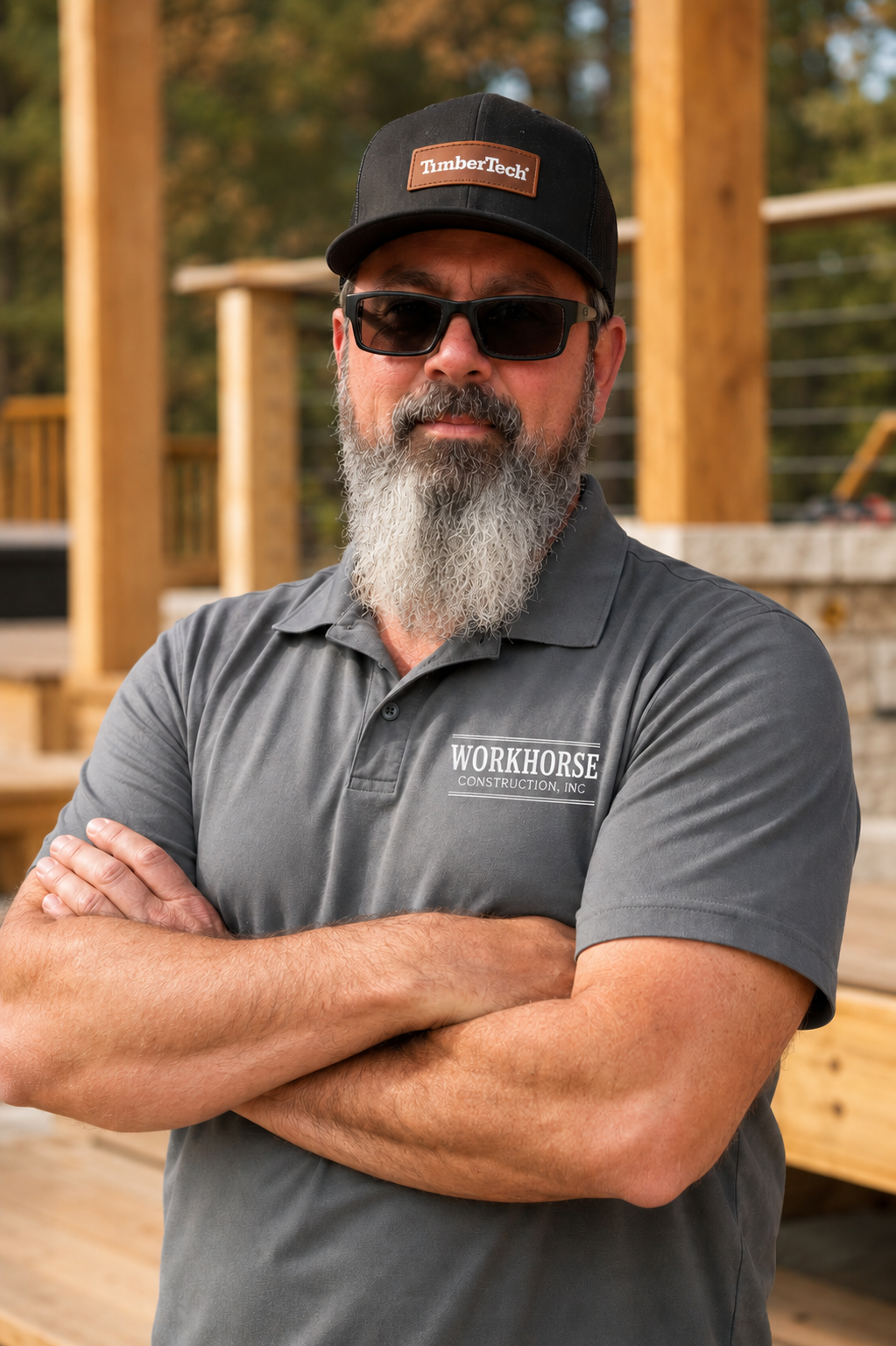 A man with a gray beard wearing sunglasses, a black cap with 'TimberTech' on it, and a gray polo shirt with 'Workhorse Construction, Inc.' on it, standing with arms crossed at a construction site with wooden framing in the background.