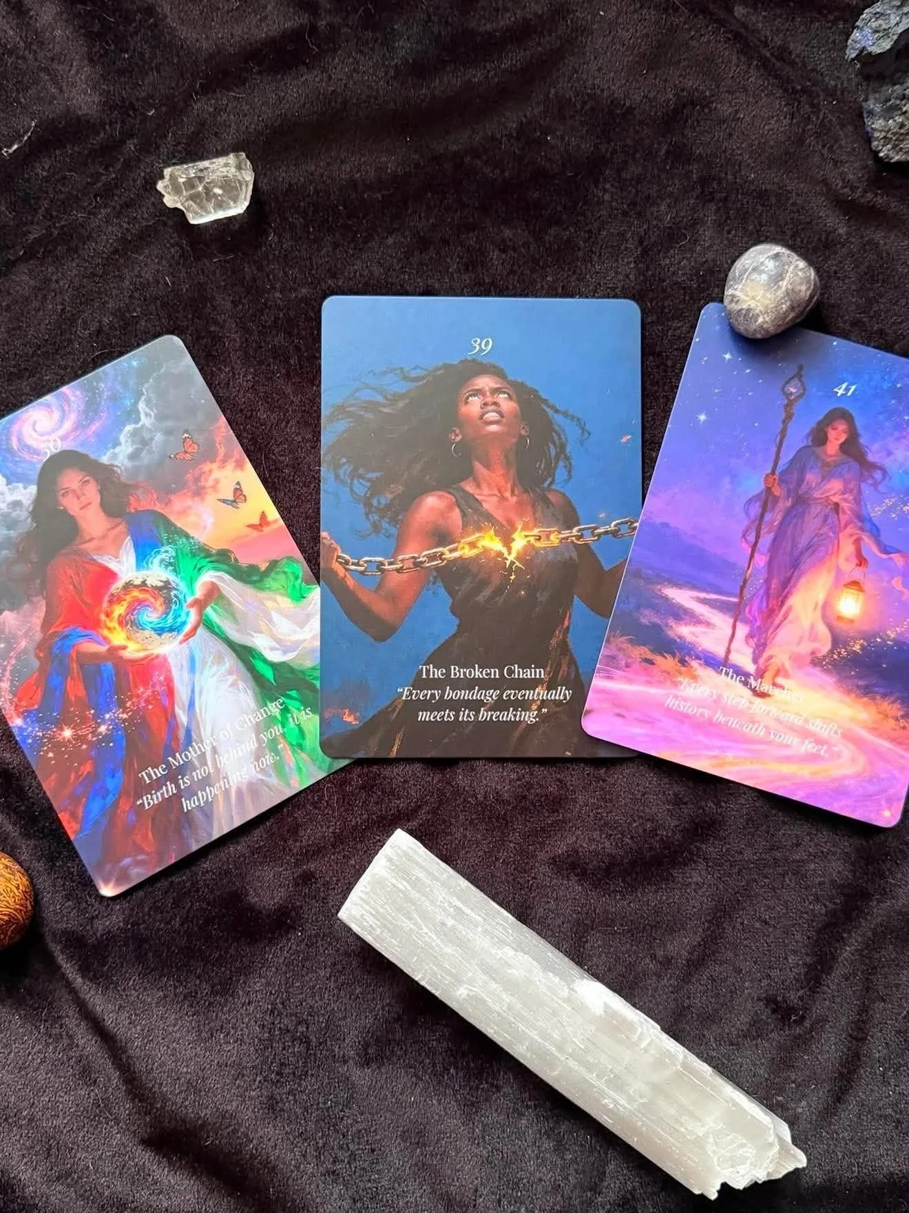 Three tarot cards on dark fabric, accompanied by a clear crystal and a small stone. The left card features a woman with a cosmic background and the quote about the birth of a maiden and new beginnings. The center card shows a woman with a chain and a quote about breaking free from bondage. The right card depicts a woman with a staff in a starry sky, with a quote about transforming your mind and fostering growth.