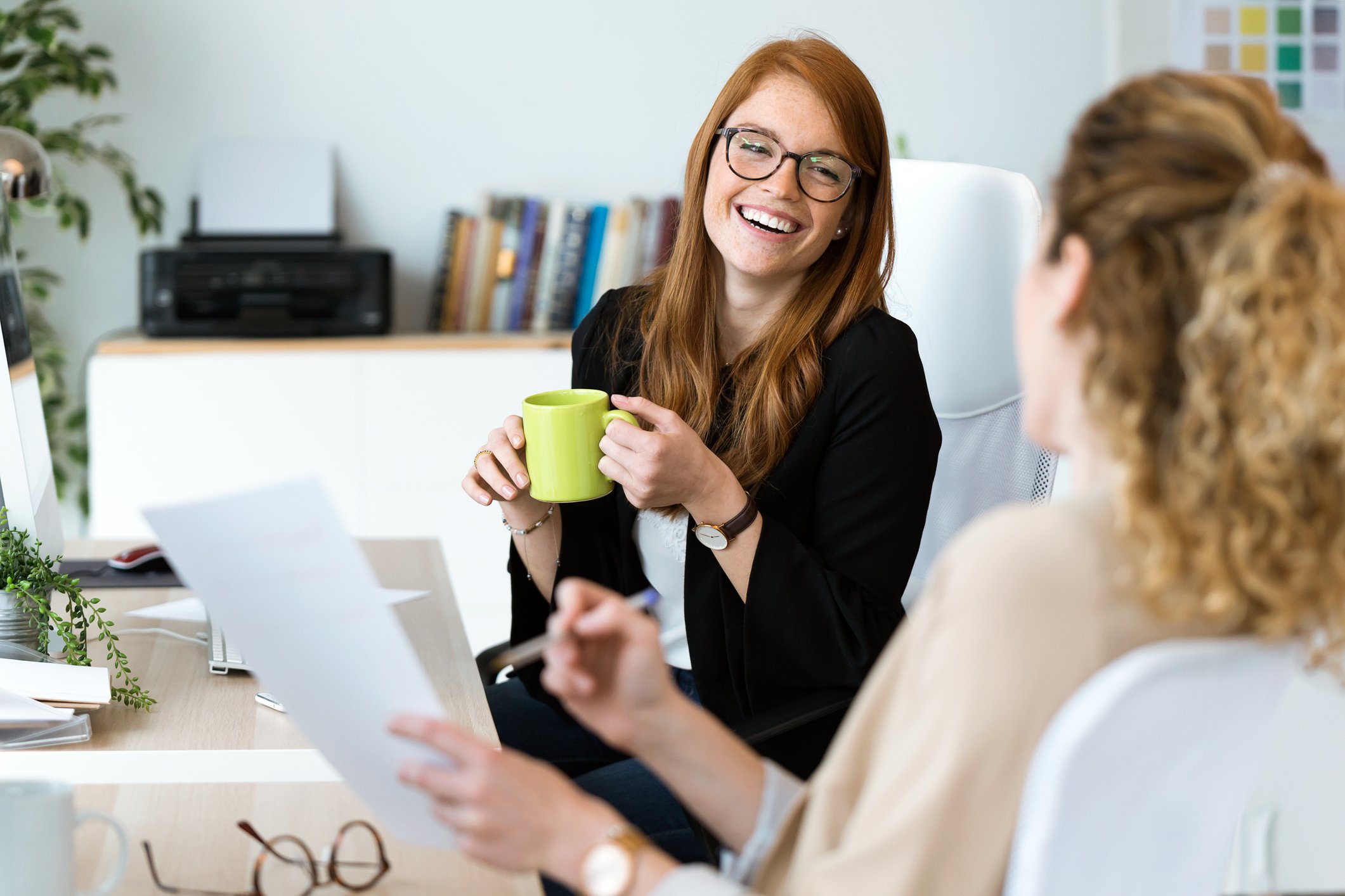 A woman with red hair, glasses, and a black blazer smiling and holding a green mug, sitting at a desk during a conversation with another woman in an office setting. Career Studio | Your next step could change everything. Practical career planning res