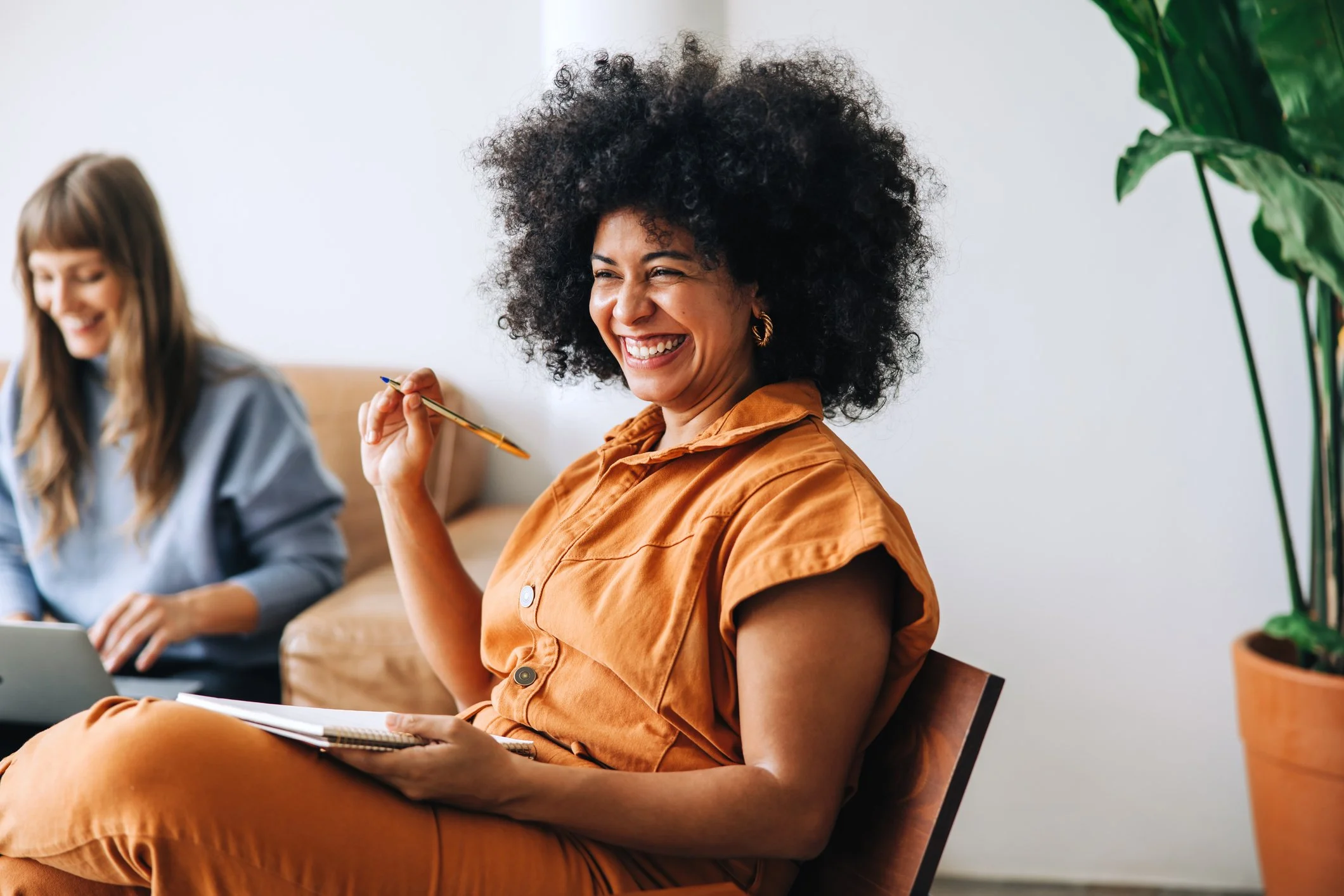 A woman with curly black hair and gold hoop earrings sitting on a chair, smiling, holding a pen and a notebook, wearing an orange shirt. Career Studio | Your next step could change everything. Practical career planning resources.