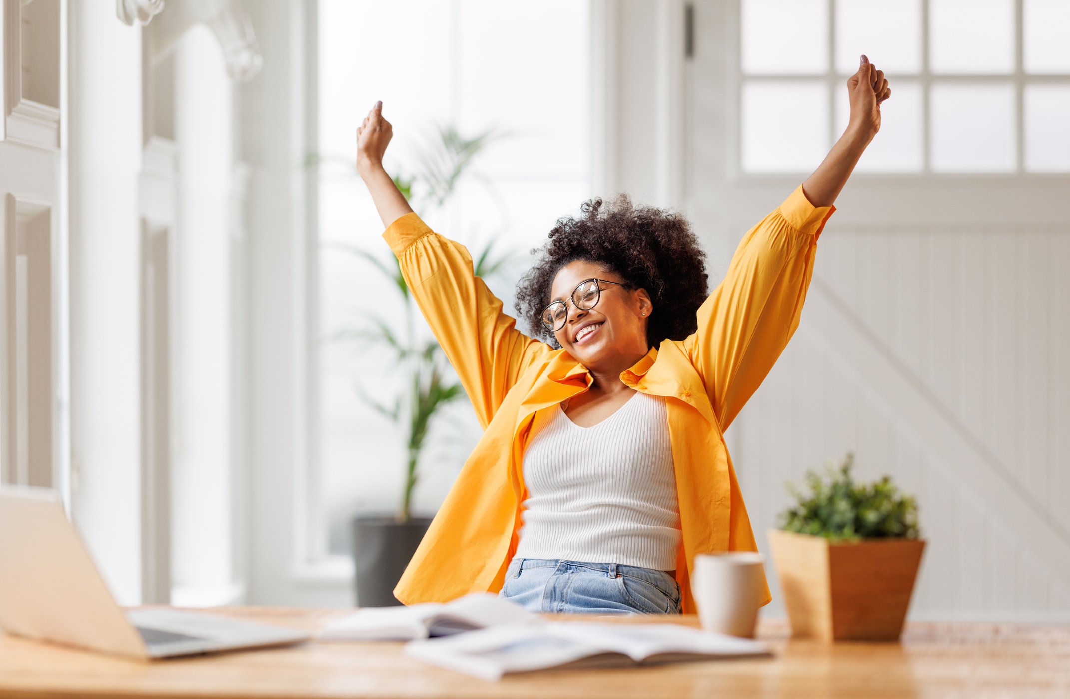 A woman with curly hair, glasses, and a big smile, stretching her arms upward while sitting at a desk in a bright room with large windows and plants. Career Studio | Your next step could change everything. Practical career planning resources.
