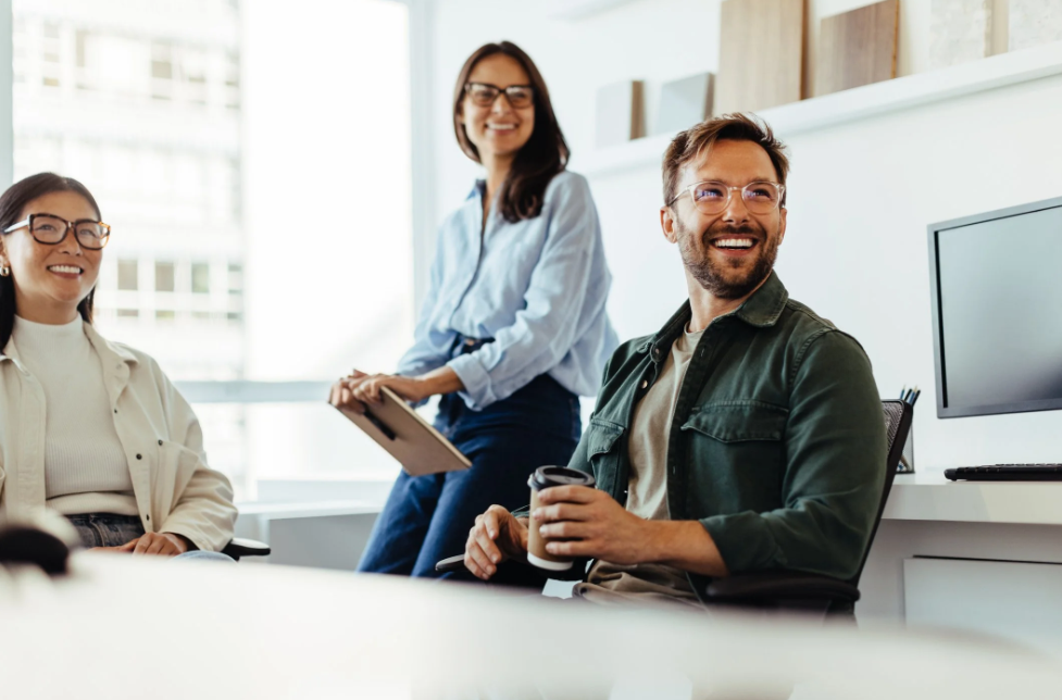 Three colleagues smiling during a meeting in a modern office, with a woman standing and holding a clipboard, and a man sitting with a coffee mug. Career Studio | Your next step could change everything. Practical career planning resources.