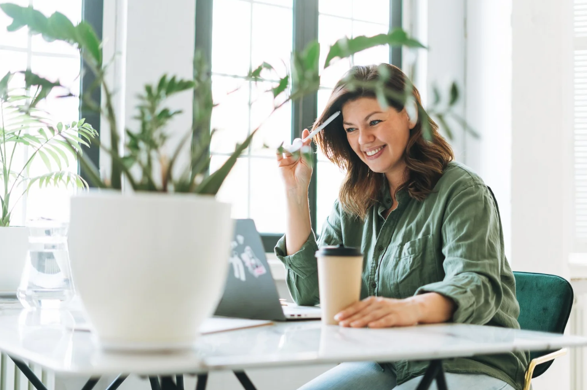 Woman with brown hair in green shirt smiling at her laptop while holding a spoon, seated at a table with a coffee cup, plants, and a bright window background. Career Studio | Your next step could change everything. Practical career planning resources