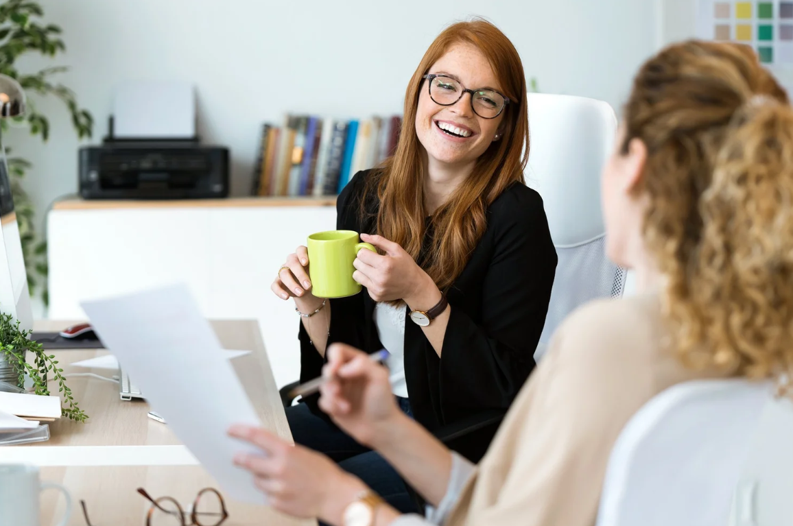 Two women having a conversation in an office, one with red hair and glasses holding a green mug and smiling, the other with curly blonde hair holding papers. Career Studio | Your next step could change everything. Practical career planning resources.