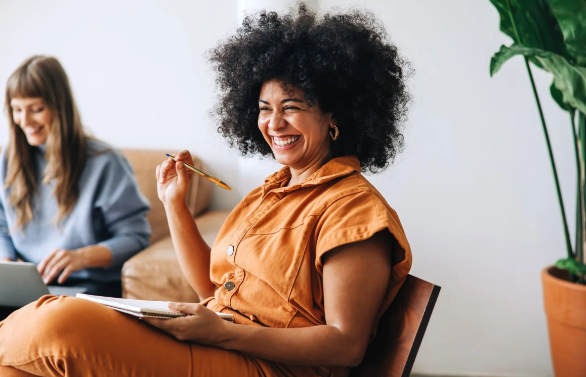 A woman with curly hair wearing an orange outfit, laughing and holding a pen and notebook, sitting in front of a woman with long hair working on a laptop, in a bright room with a potted plant. Career Studio | Your next step could change everything. P