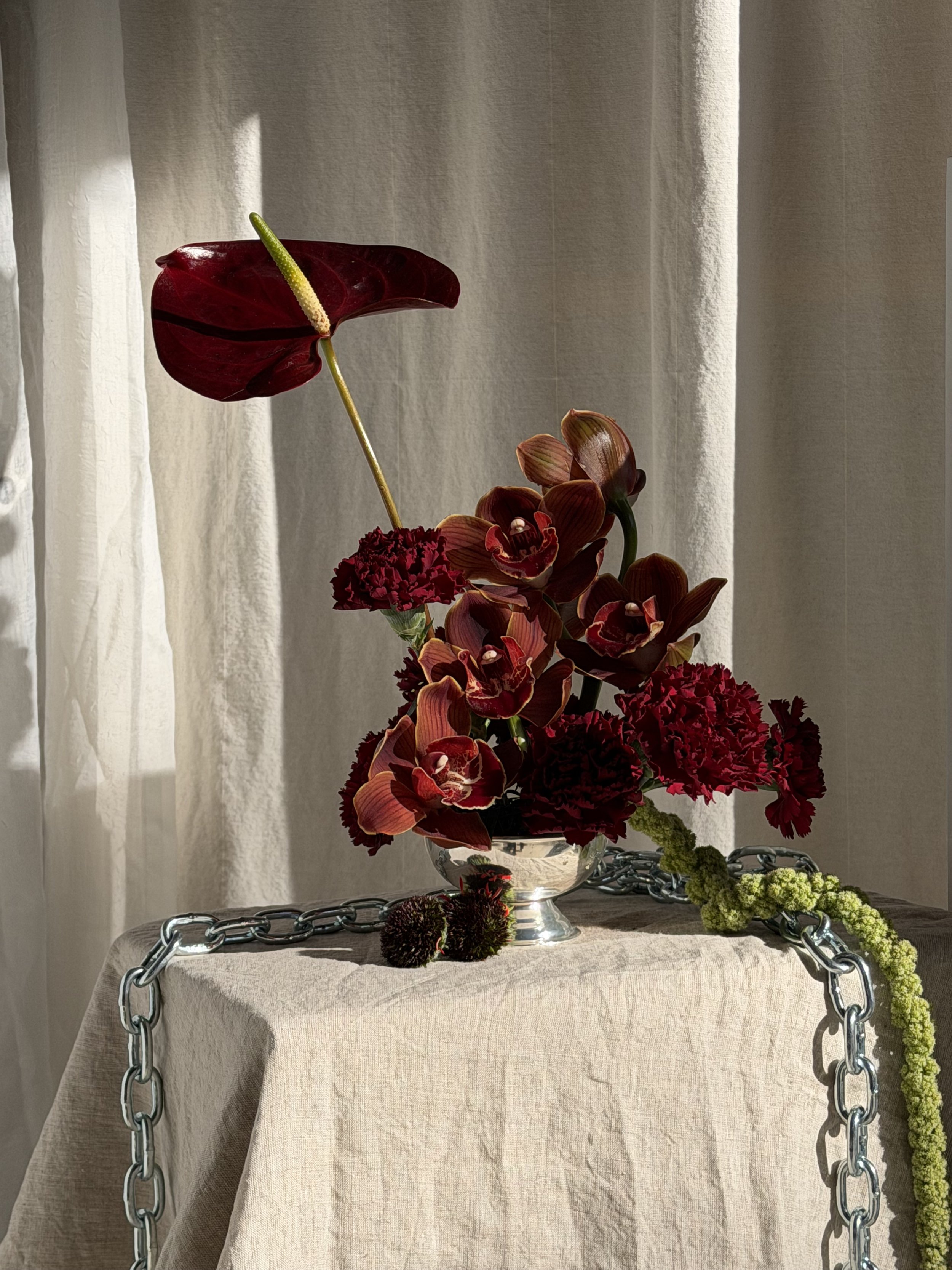 A floral arrangement with red anthuriums, maroon orchids, and red carnations in a silver vase on a table draped with beige cloth, adorned with a chain around the table, with curtains and sunlight in the background.