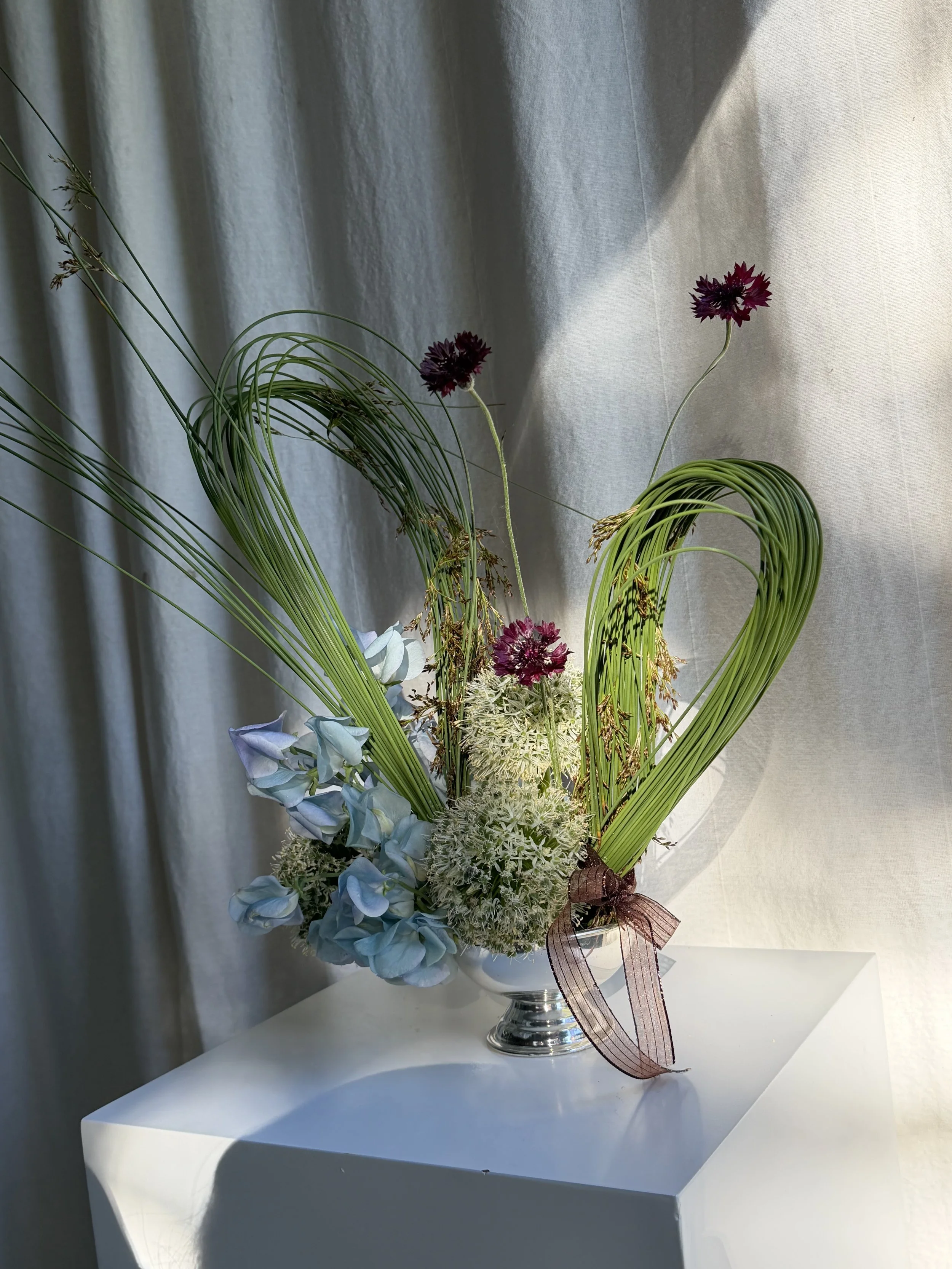 A floral arrangement in a silver vase with purple, white, and blue flowers and green foliage, set on a white table against a light-colored curtain background.