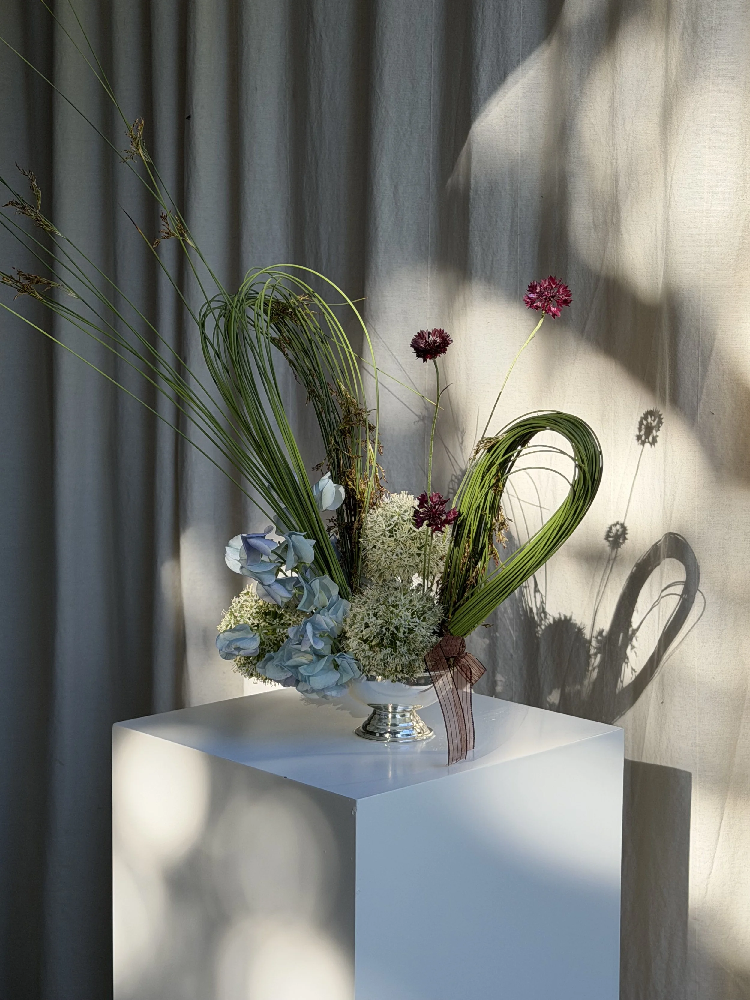 A floral arrangement in a silver vase with pink, light blue, and white flowers, placed on a white pedestal against a curtain backdrop, with shadows cast on the curtain.