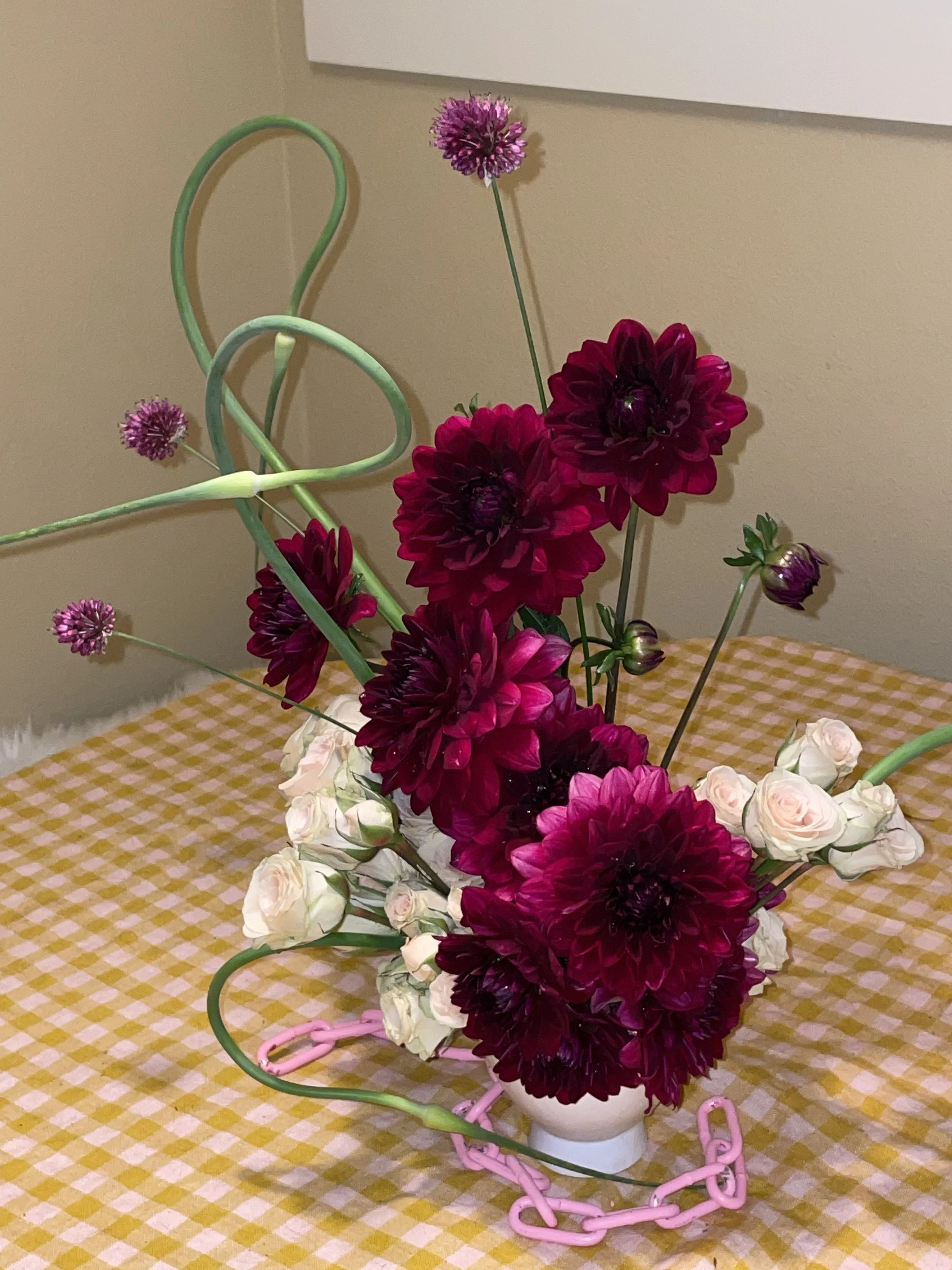 A floral arrangement in a white vase with purple, maroon, and cream flowers, placed on a table with a yellow checkered tablecloth and pink chain decorations.