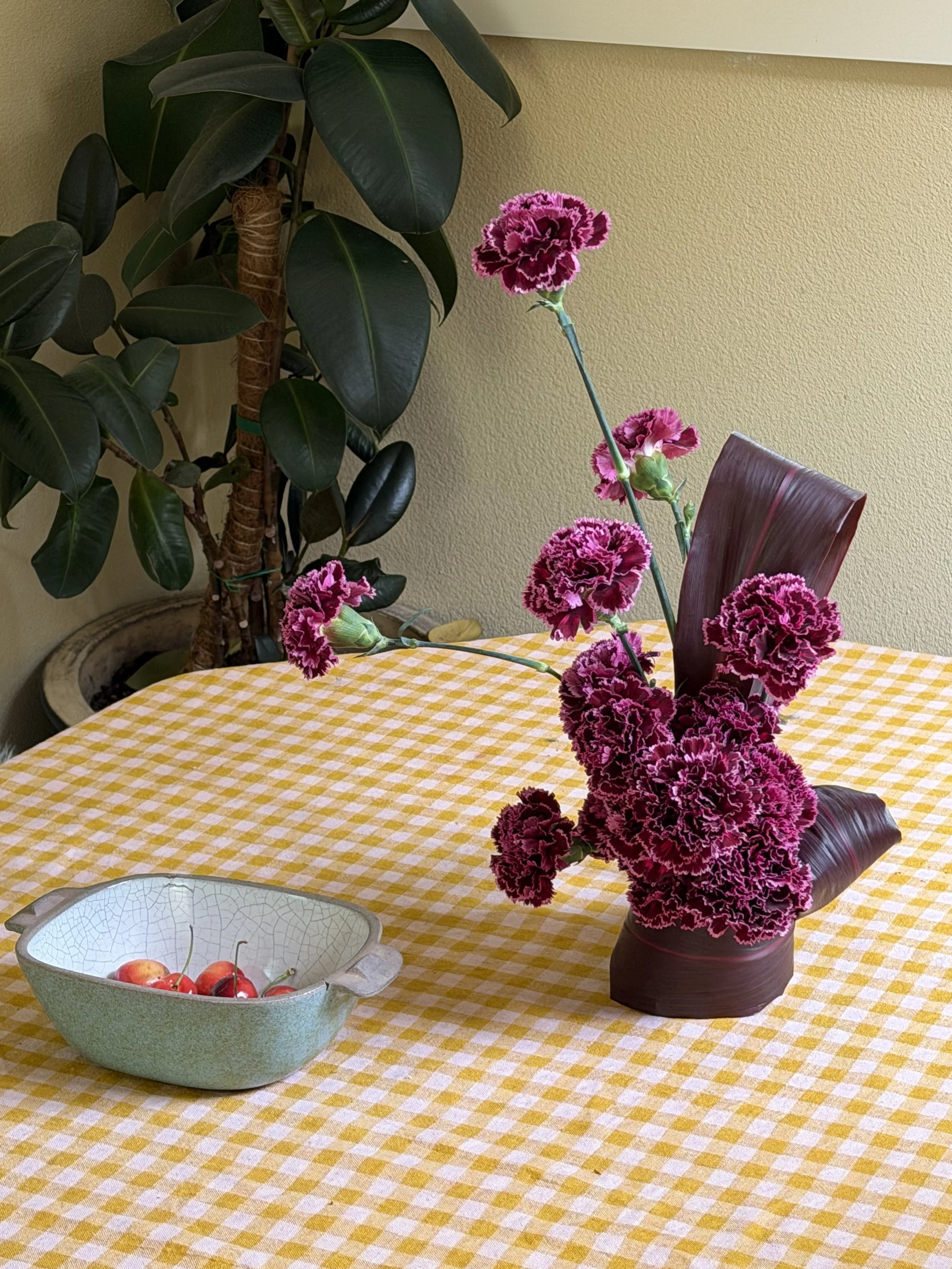 A pink and purple carnation flower arrangement in a dark purple vase on a yellow checkered tablecloth, with a green potted plant with large dark green leaves in the background, and a bowl of cherries on the table.