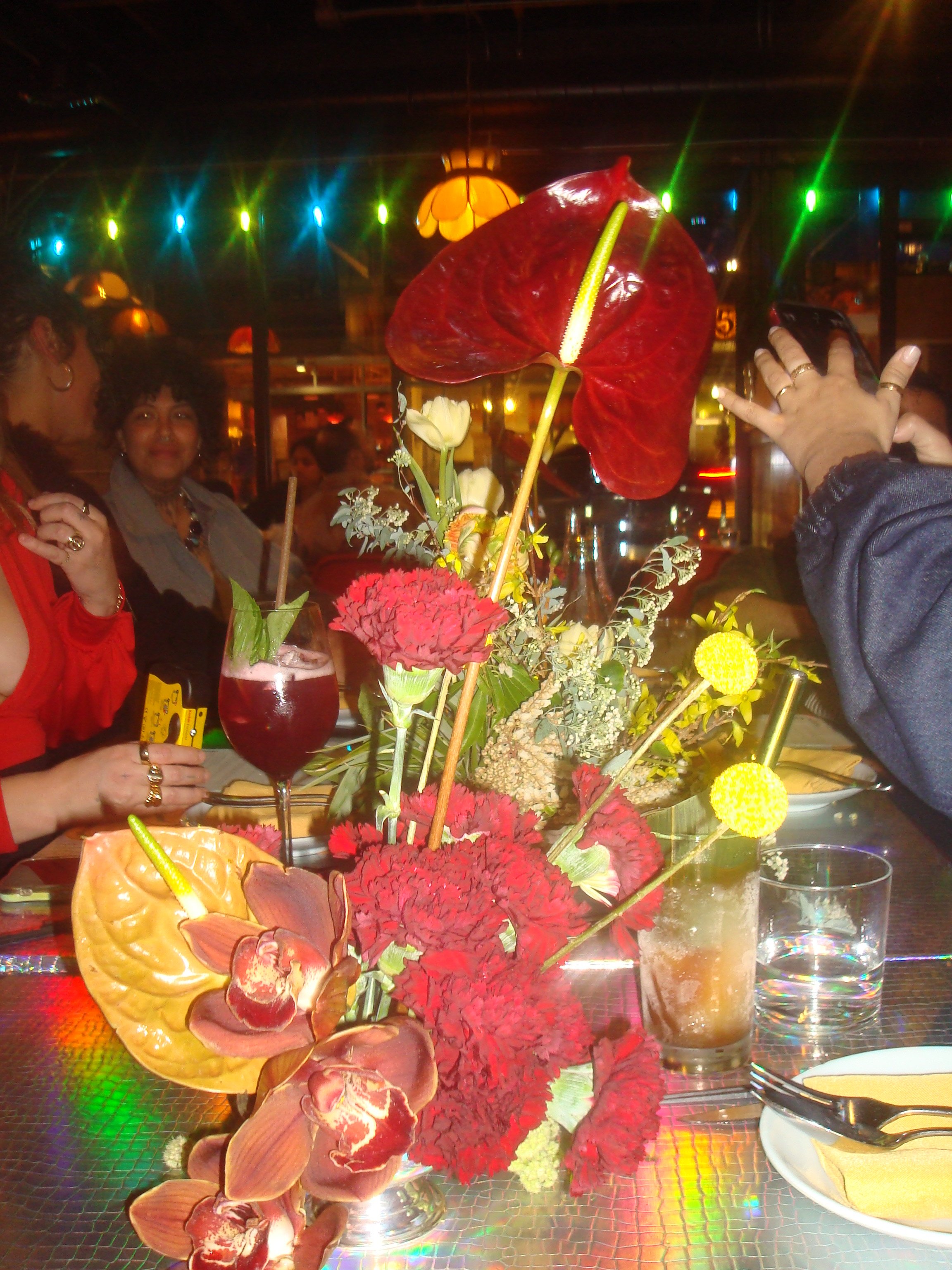A restaurant table with a colorful floral centerpiece, drinks, and diners in the background under neon lights.