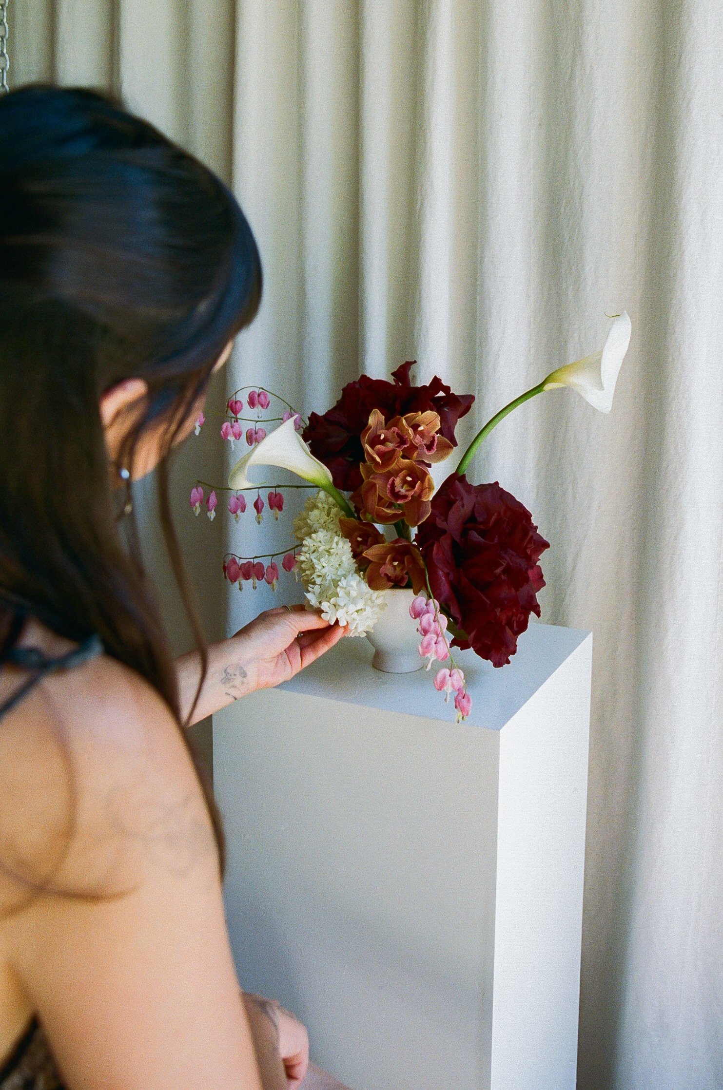 A woman is arranging a floral bouquet on a white pedestal in front of a light-colored curtain.