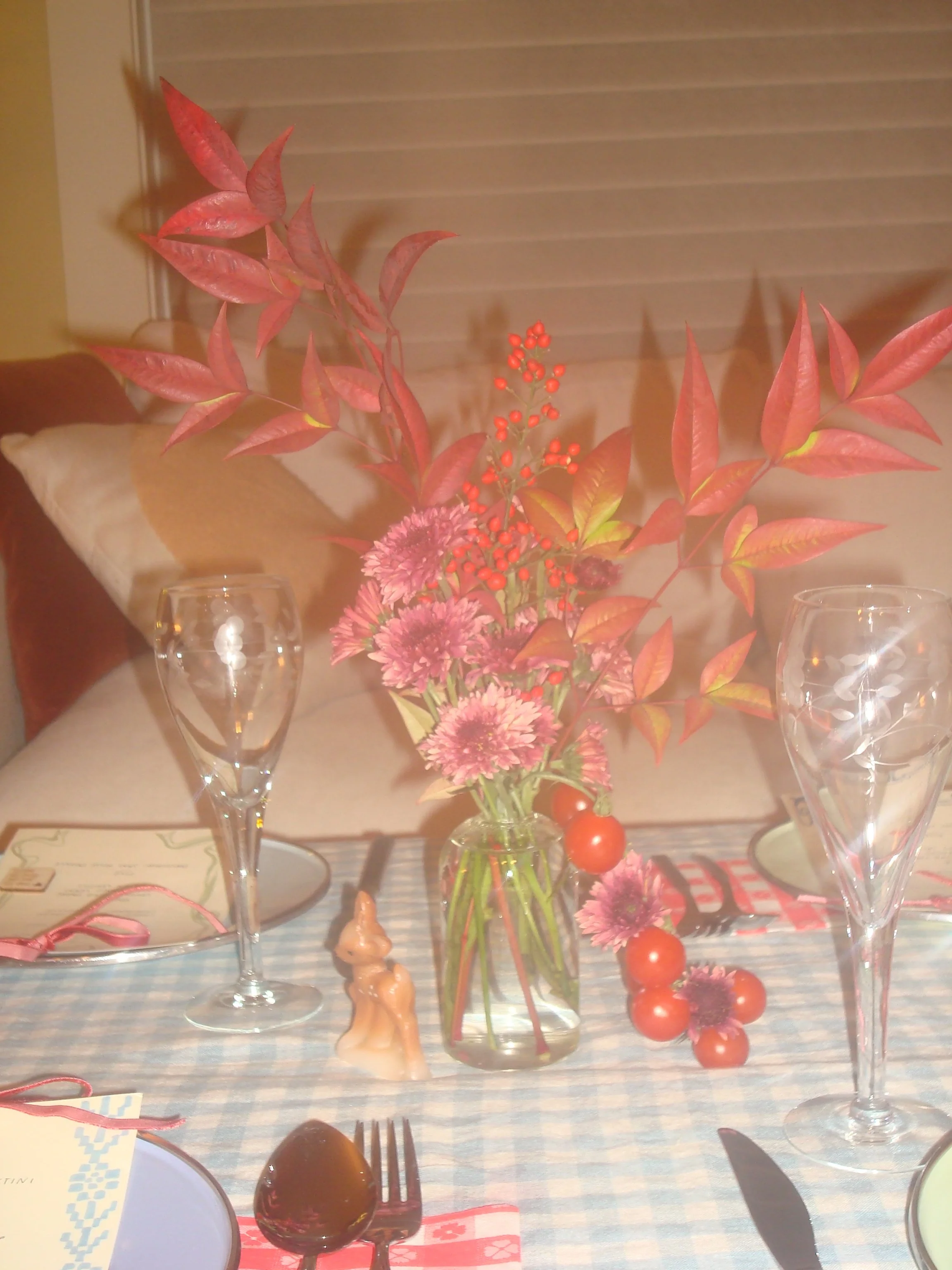 A table with a floral centerpiece in a glass vase, two empty wine glasses, a chocolate egg with a fork, a paper plate with a blue pattern, and some holiday decorations. The background includes a window with blinds and a cushion.