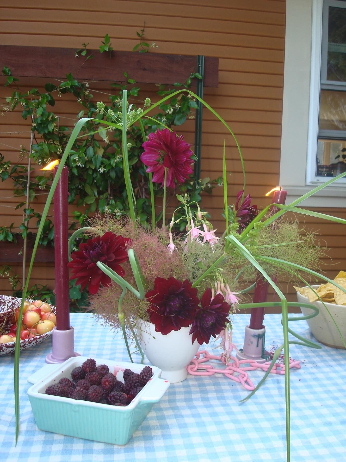 A table with a blue checkered tablecloth features a white vase with pink and red flowers, surrounded by pink candle holders, with bowls of cherries and blackberries nearby, set against a wooden wall and window background.