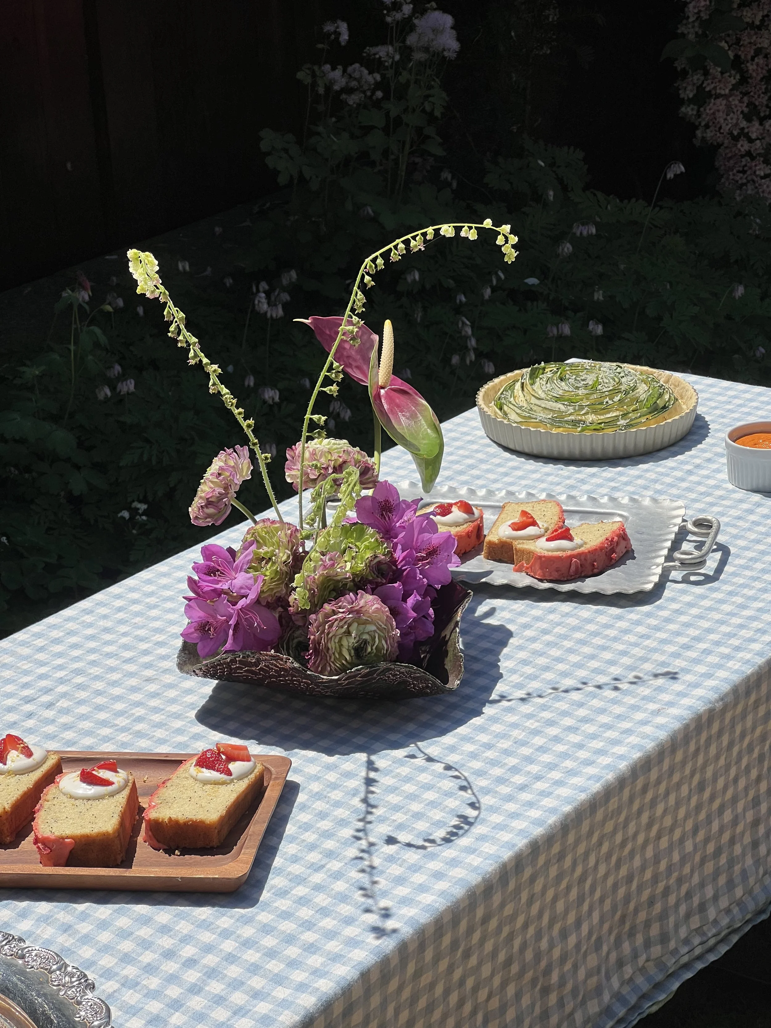 A table with a blue and white checkered tablecloth displaying a floral arrangement in a vessel, slices of cake with strawberries and whipped cream, and a green vegetable pie in a white dish, set outdoors at night.