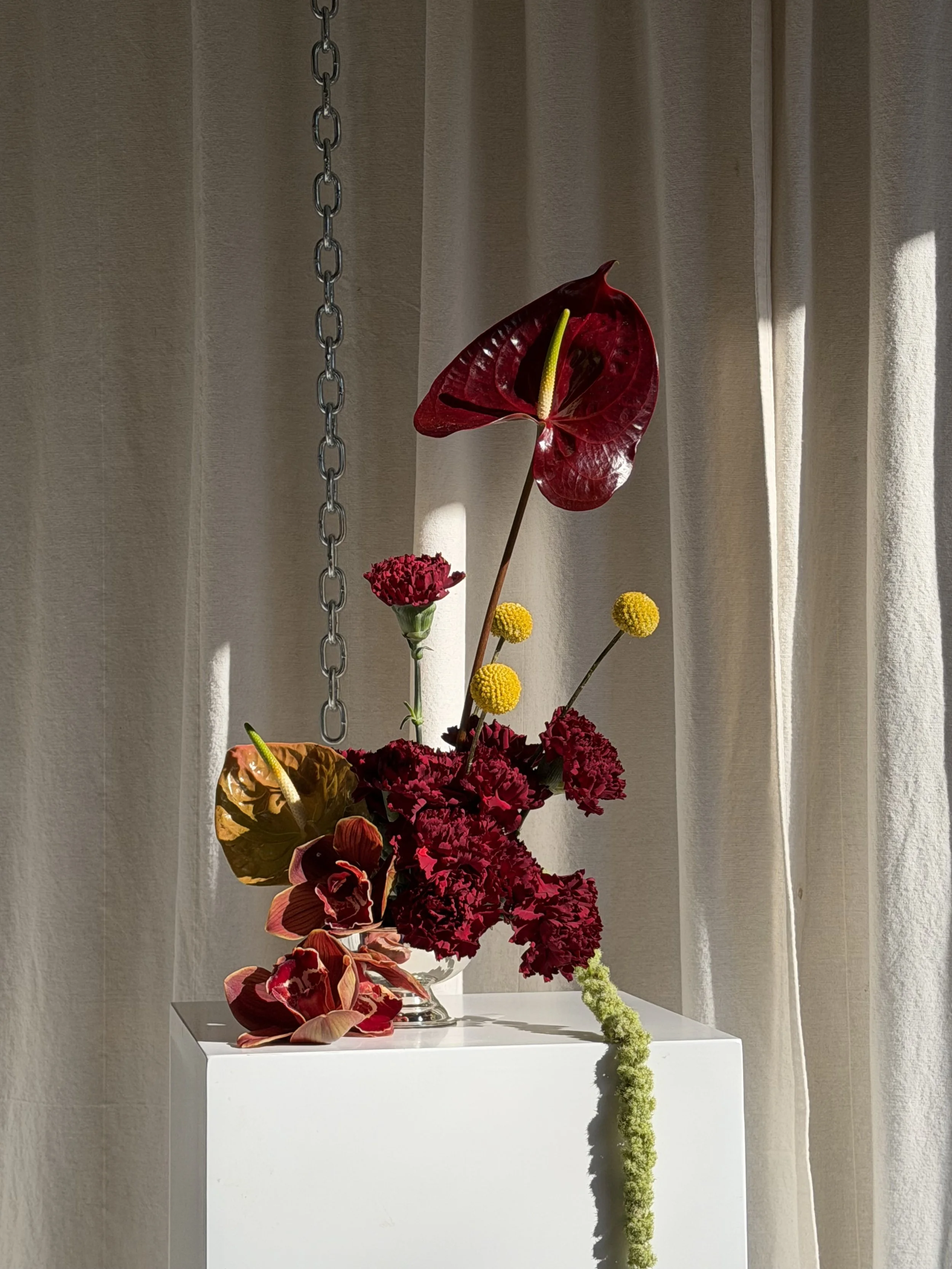 A floral arrangement with red anthuriums, pink carnations, yellow billy balls, and a trailing greenish-yellow flower, displayed on a white pedestal with beige curtains in the background.