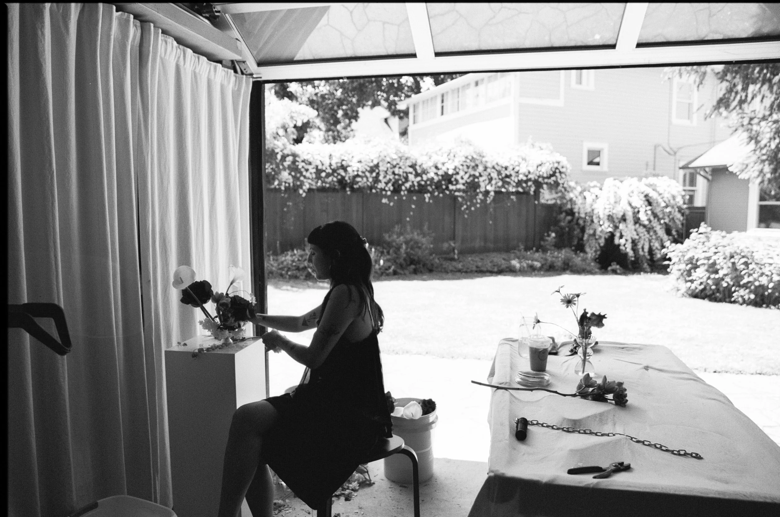 A woman with long dark hair sitting on a stool indoors, with her profile visible, holding a bouquet of flowers. She is near a table with a white tablecloth, decorated with more flowers and some objects. Large windows behind her reveal an outdoor gard