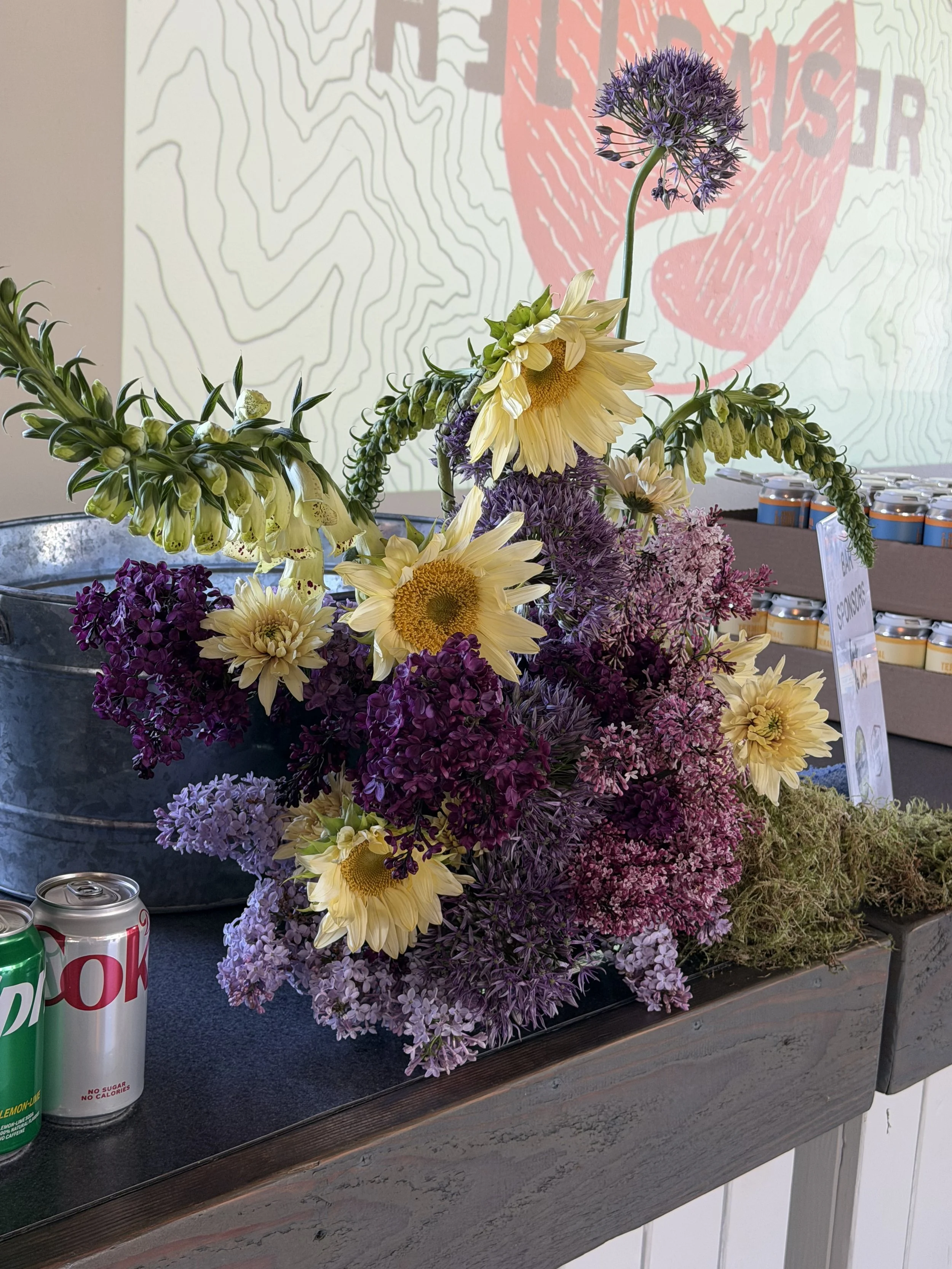 A floral arrangement with yellow sunflowers, purple alliums, and other purple and pink flowers on a black table, with soda cans nearby and a red and white sign behind it.