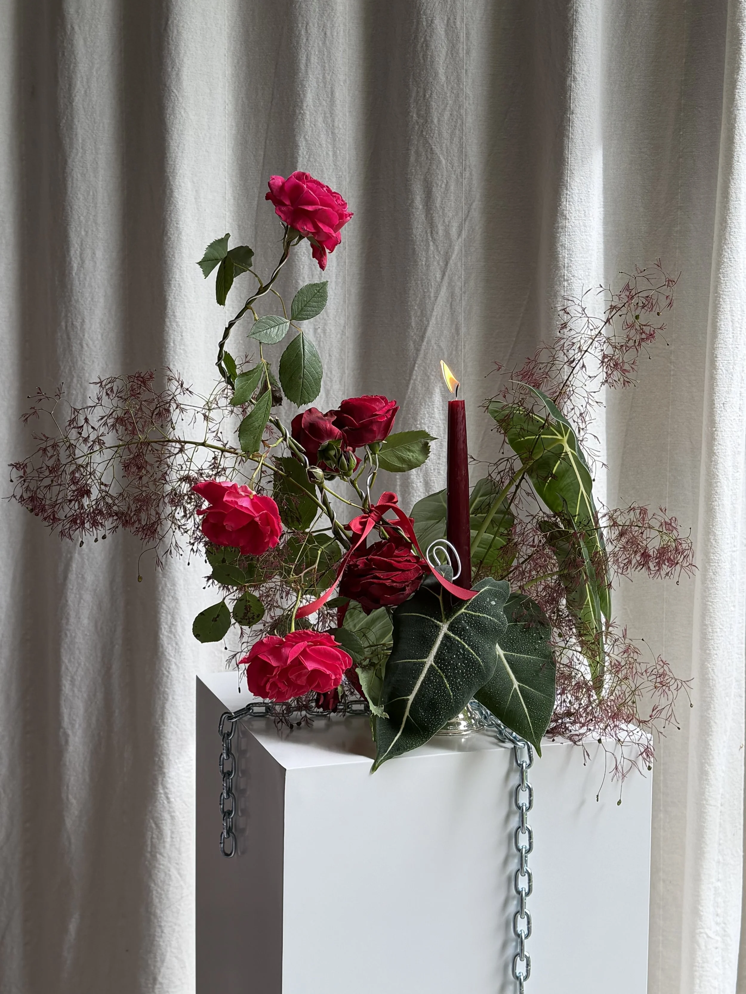 A bouquet of red roses, green leaves, and thin, red, branching flowers arranged with a lit red candle on a white pedestal with chains hanging from the sides, in front of a beige curtain.