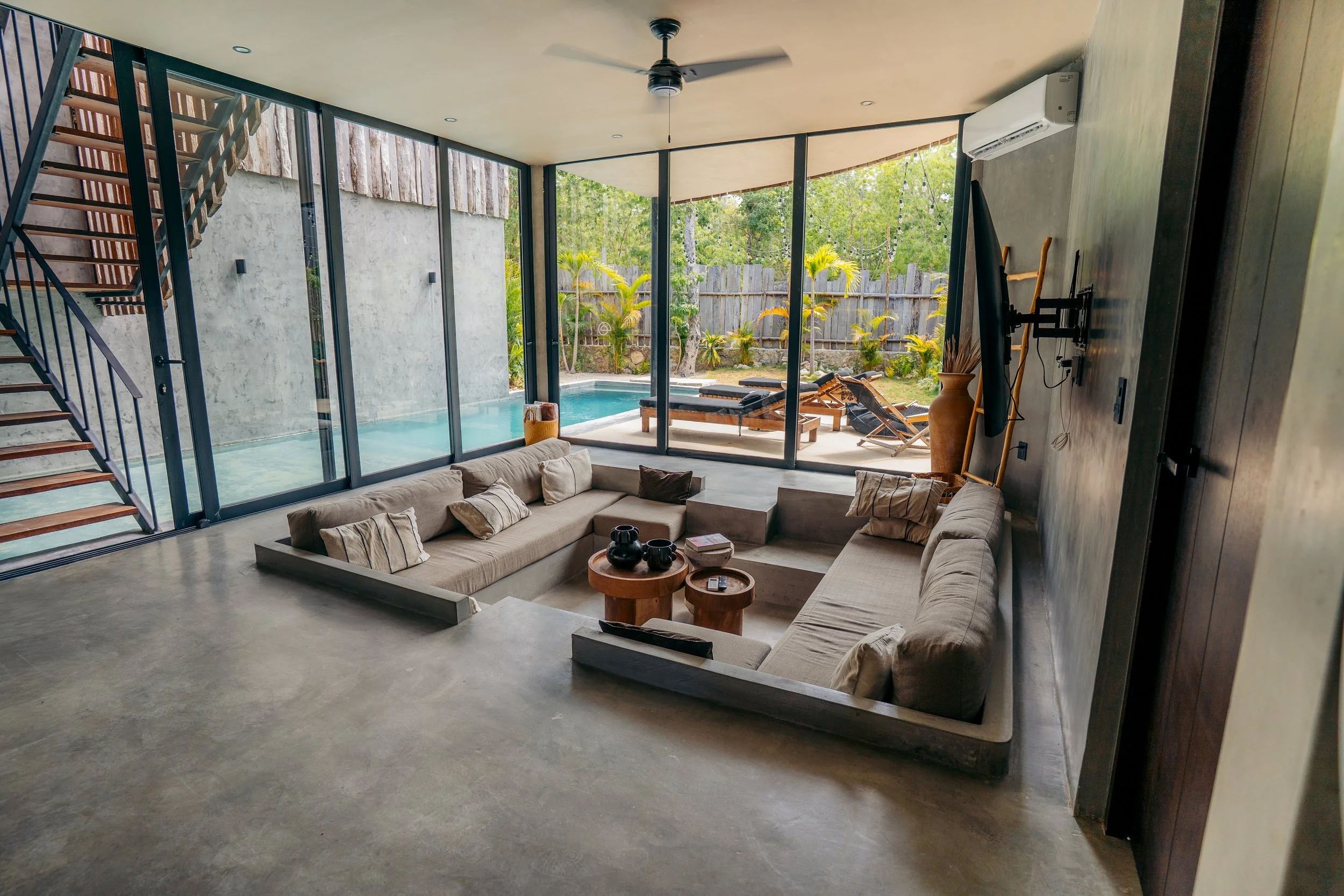 Living room with large glass doors opening to a backyard pool area, featuring outdoor lounge chairs, a wooden fence, and lush green plants.