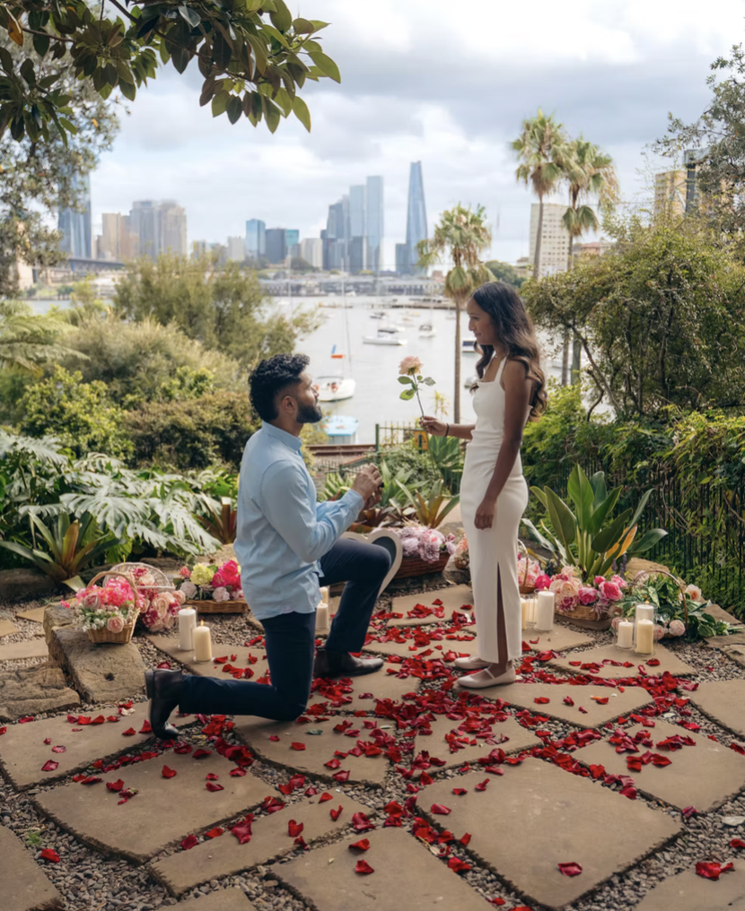 A man proposes to a woman on a rose petal covered pathway adorned with candles and flowers, overlooking a city skyline and a waterfront with boats, under a partly cloudy sky.