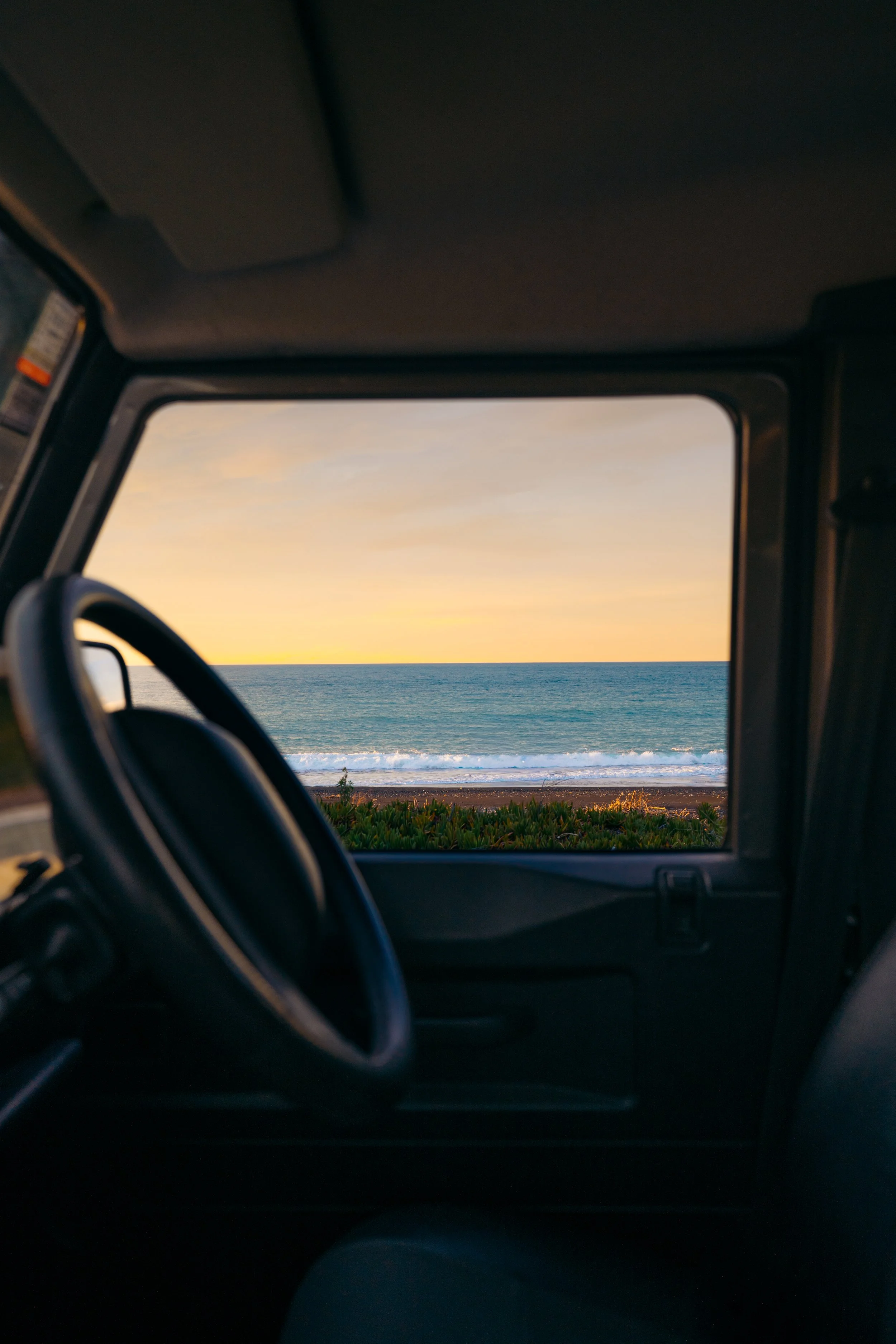 Inside a vehicle showing the front dashboard and steering wheel, with a scenic view of the ocean and a sunset through the open window.