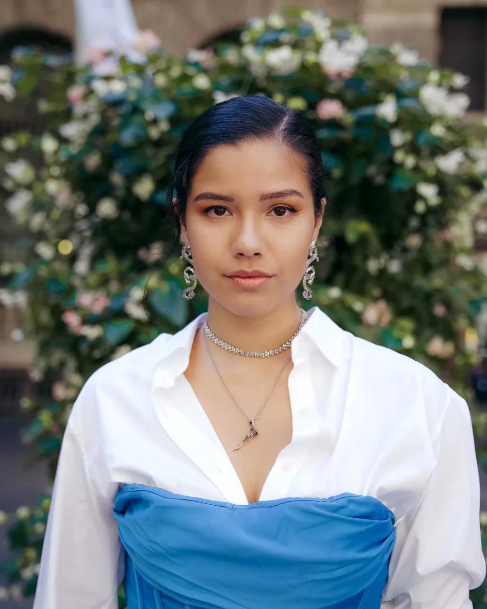 A woman with dark hair styled back, wearing statement earrings, layered necklaces, a white shirt, and a blue strapless dress, standing outdoors in front of lush greenery with pink and white flowers.