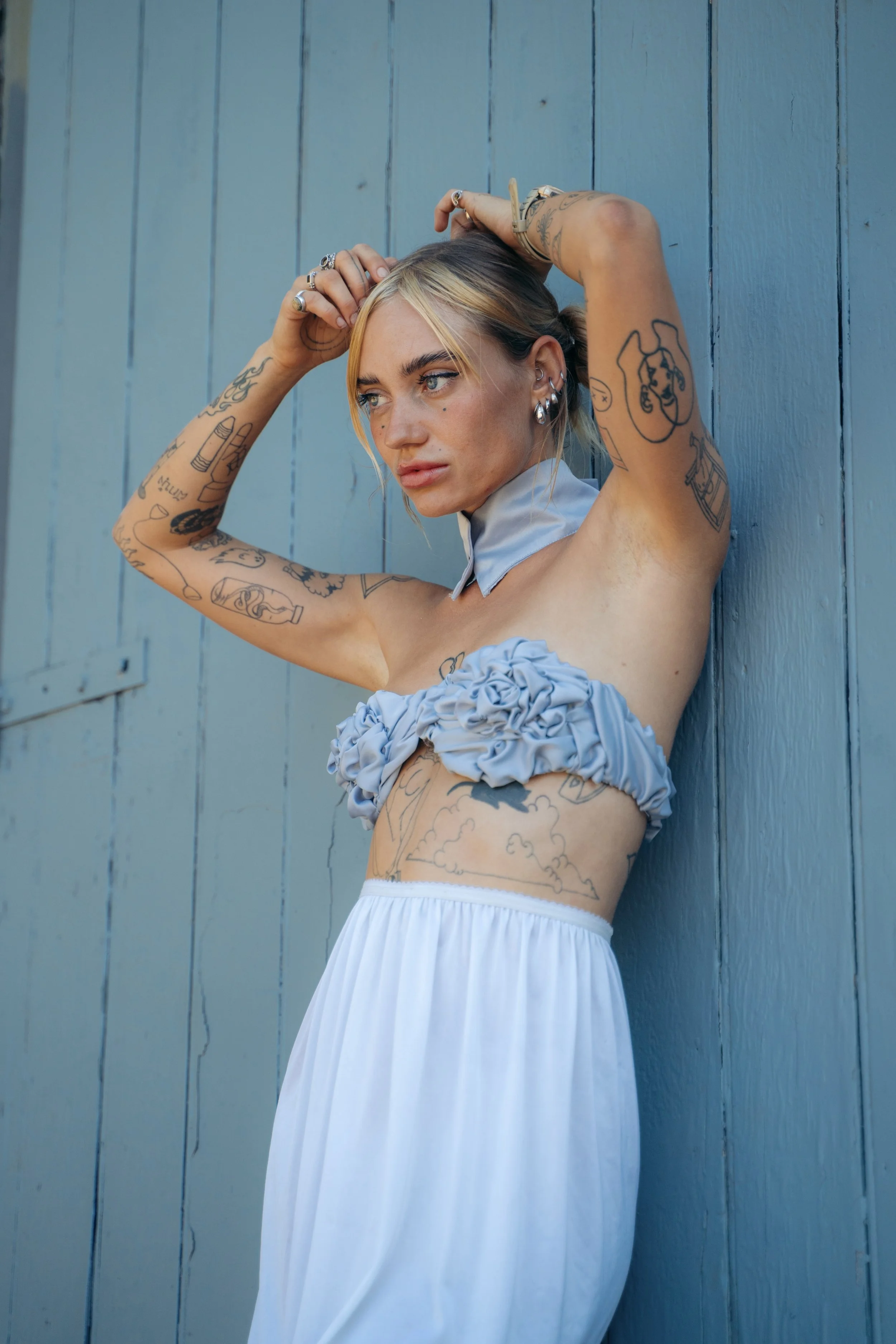 A young woman with tattoos and multiple earrings, wearing a light blue off-shoulder dress, poses against a blue wooden wall, looking to her left with a serious expression.