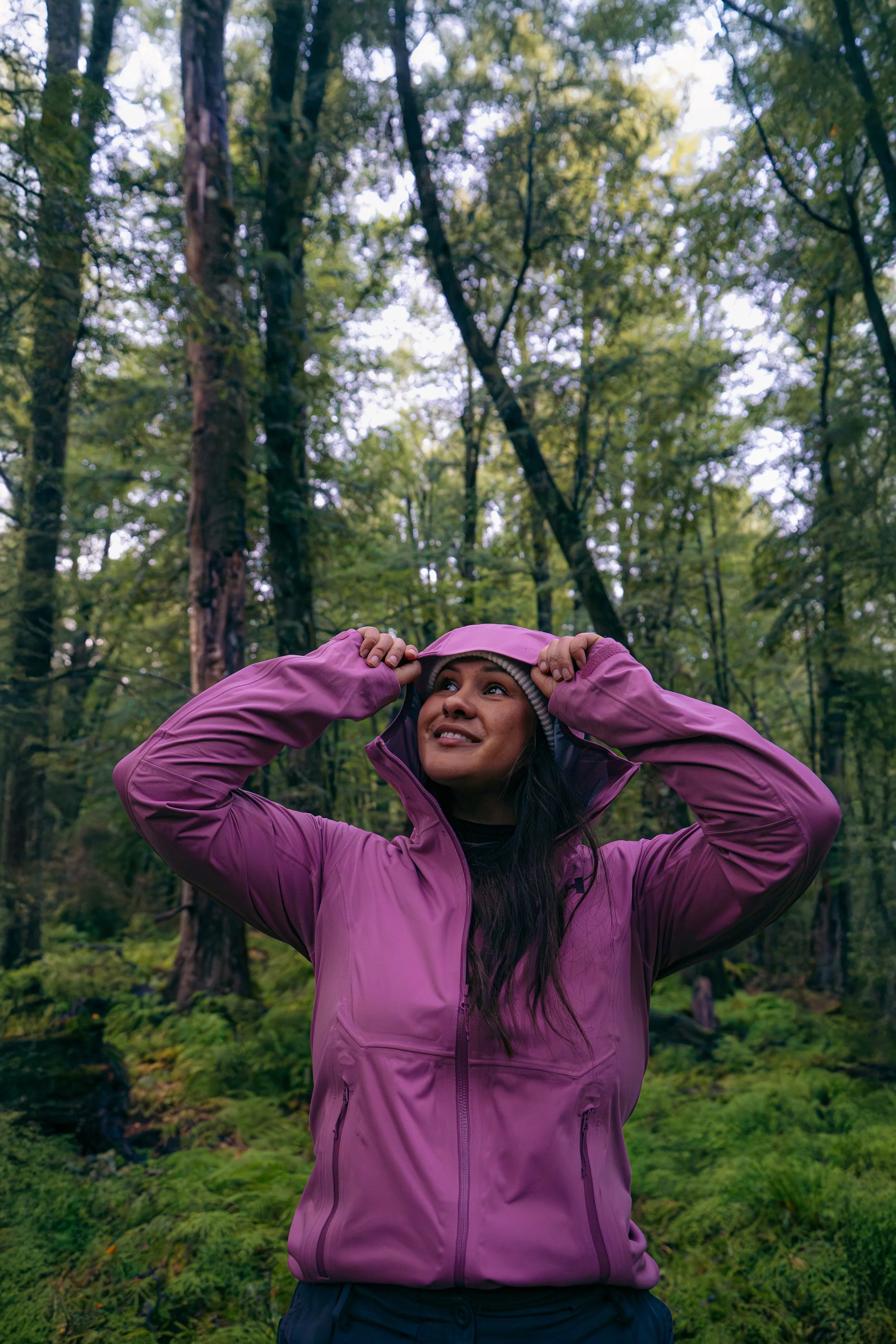 A woman in a pink outdoor jacket and gray beanie adjusting her hood in a forest with tall trees and green foliage.