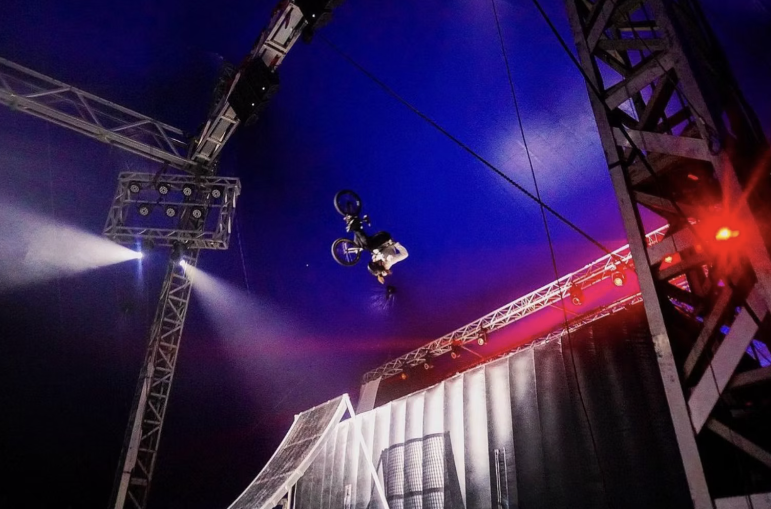 A BMX rider performing an aerial stunt at a nighttime indoor event, with stage lights and metal scaffolding structures around.